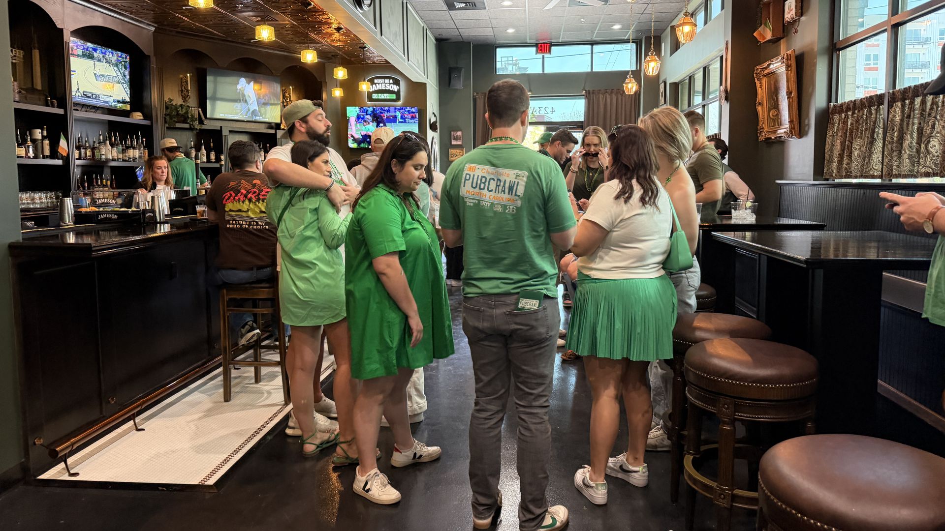 Inside a bustling bar, people in green chat near the black bar. Bottles line the left, TVs show sports, warm pendant lights glow, and large windows brighten the right side.