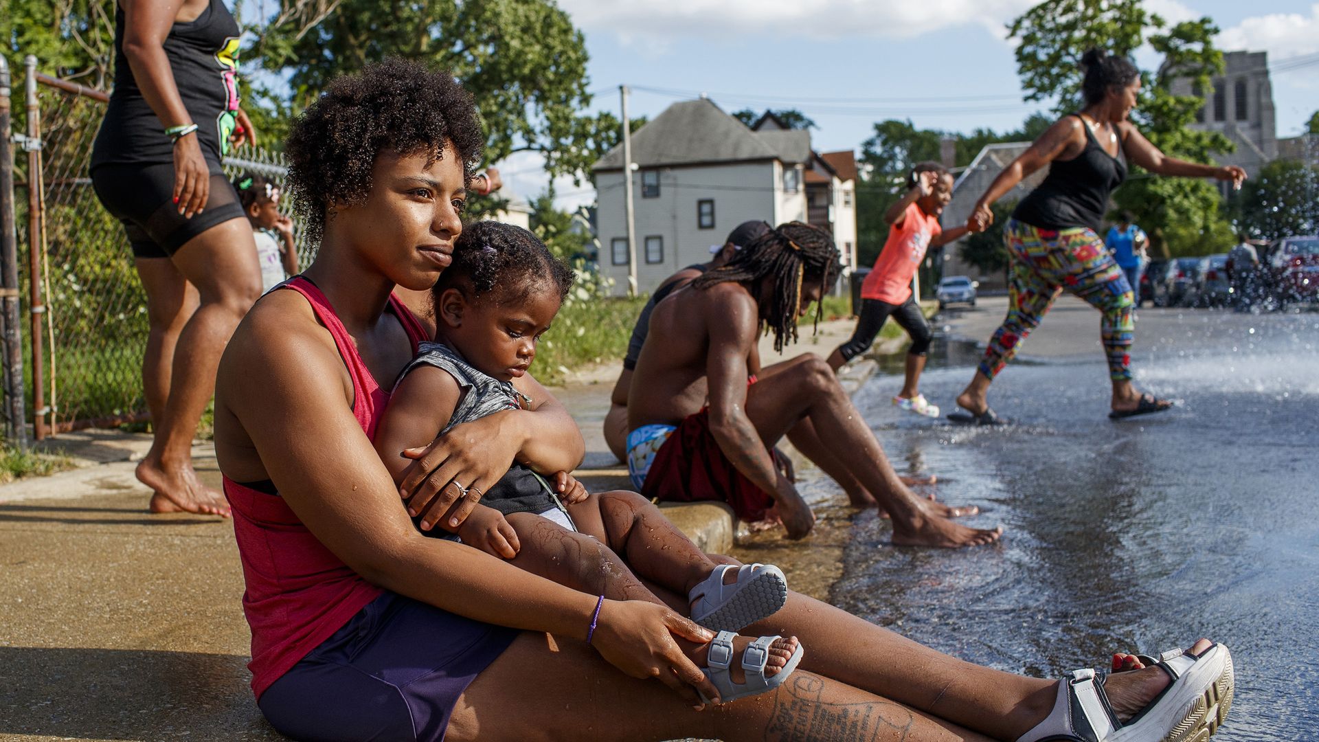 A woman sits on the sidewalk of a street on a hot summer day, with kids playing in spraying water in the background.