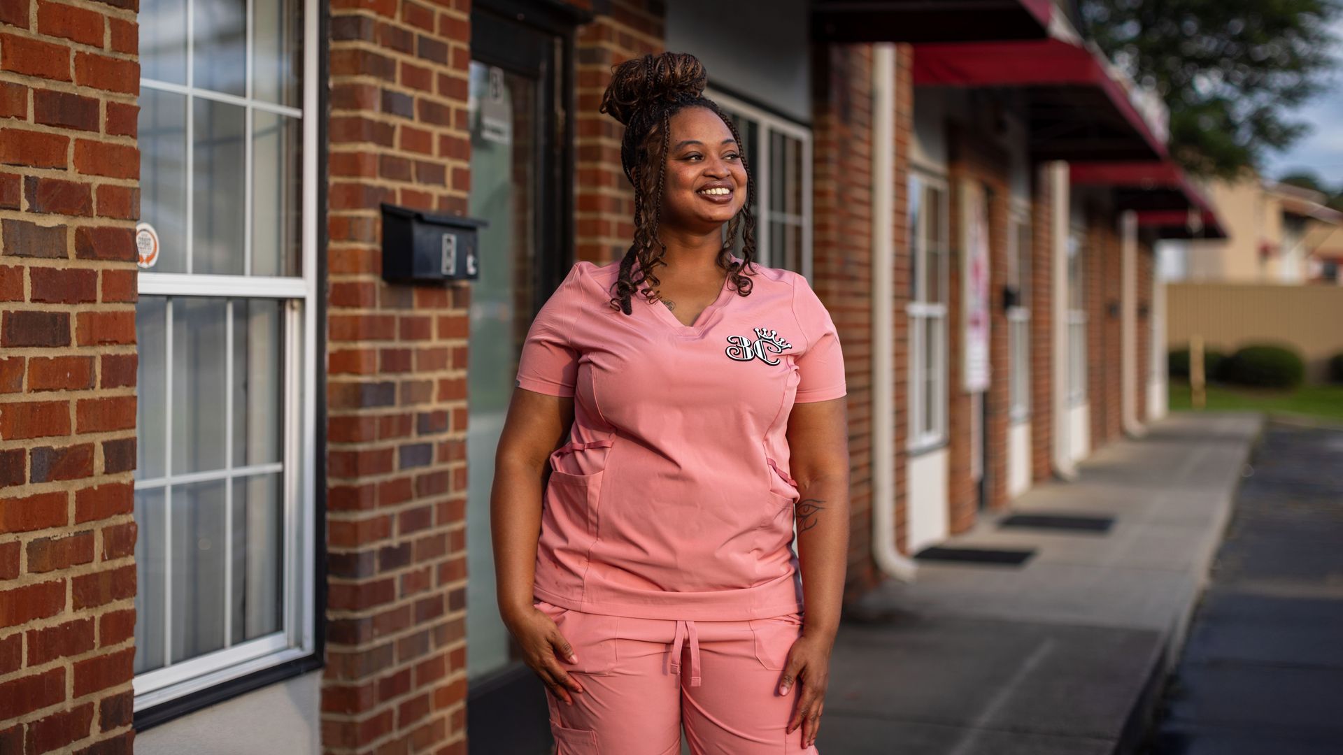 Smiling woman with braided hair in pink scrubs with a logo stands outside a brick storefront row with windows and red awnings; trees visible in distance.