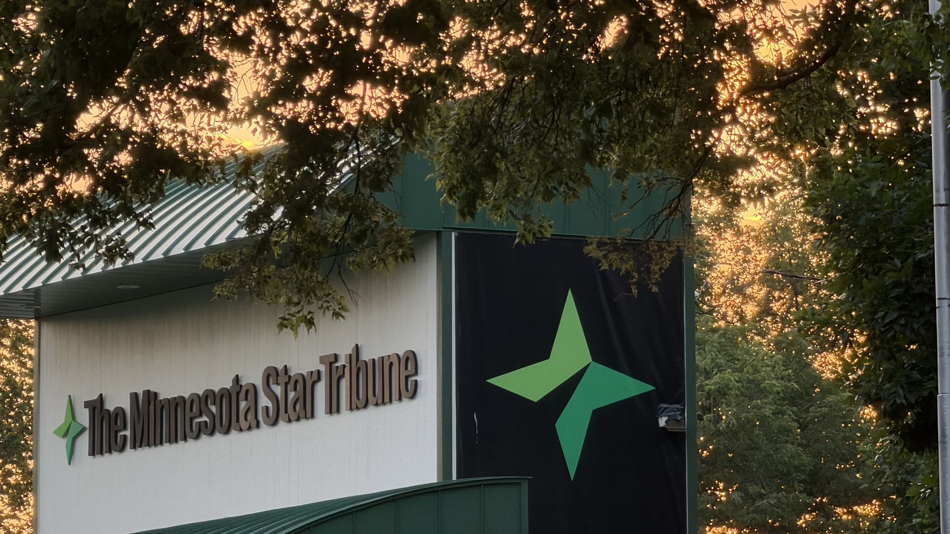 Building sign reads "The Minnesota Star Tribune" with a green star logo, green awning, and orange sunset filtering through tree branches overhead.