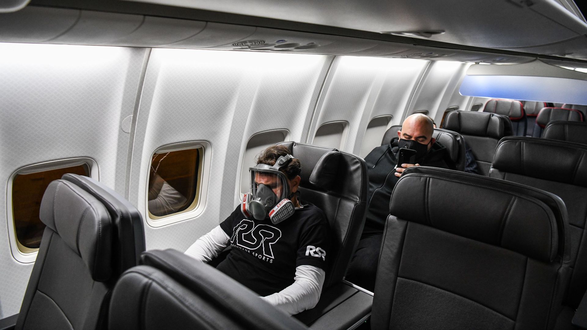 In this image, a man wears a gas mask while sitting on a plane