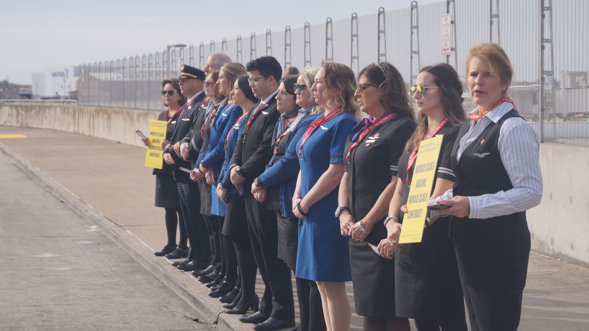Flight attendants standing in line on sidewalk near CLE, holding yellow protest signs.