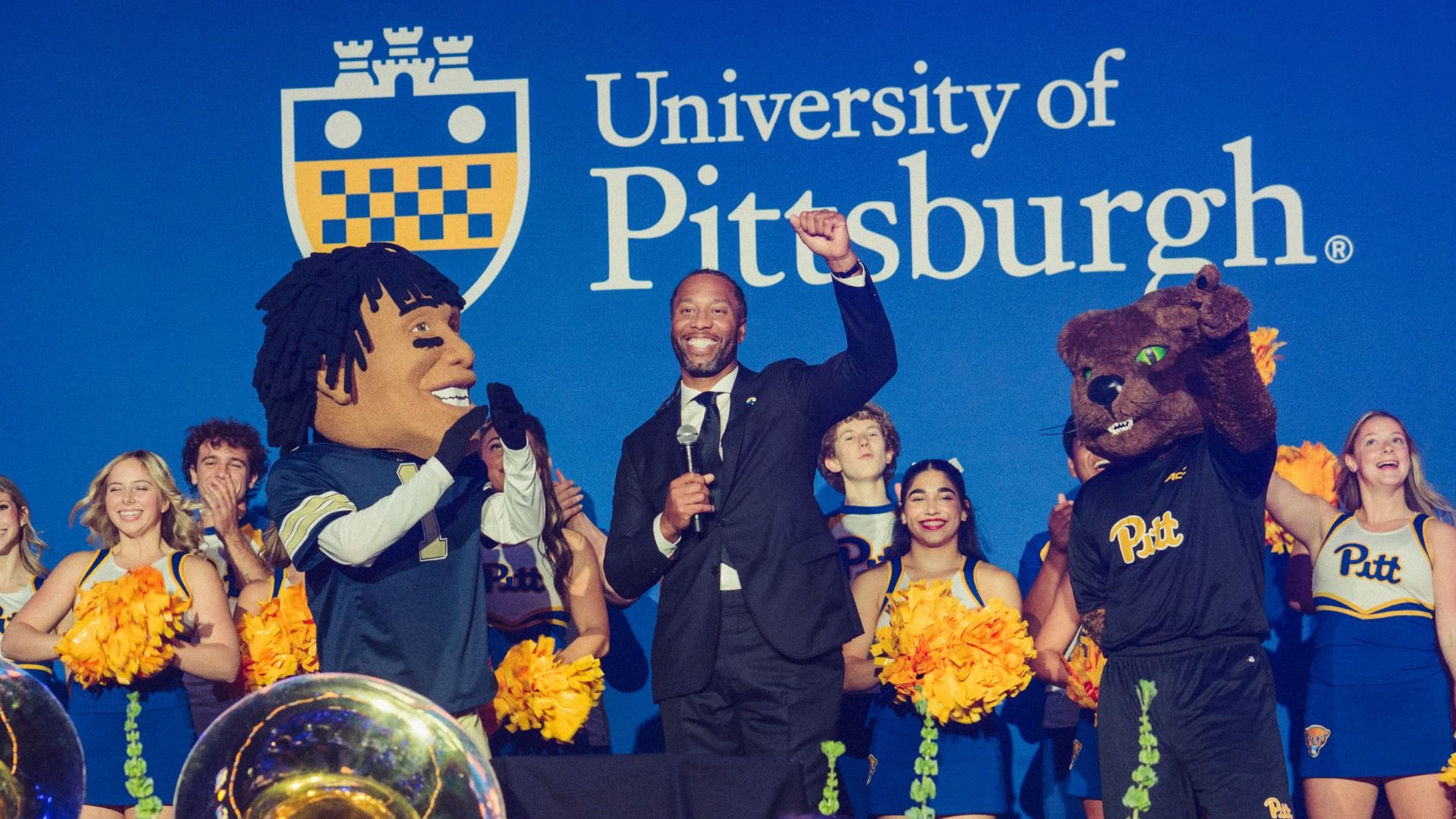 On a blue stage with the "University of Pittsburgh" sign, a man in a suit smiles and raises a fist as Pitt mascots and cheerleaders cheer beside him.