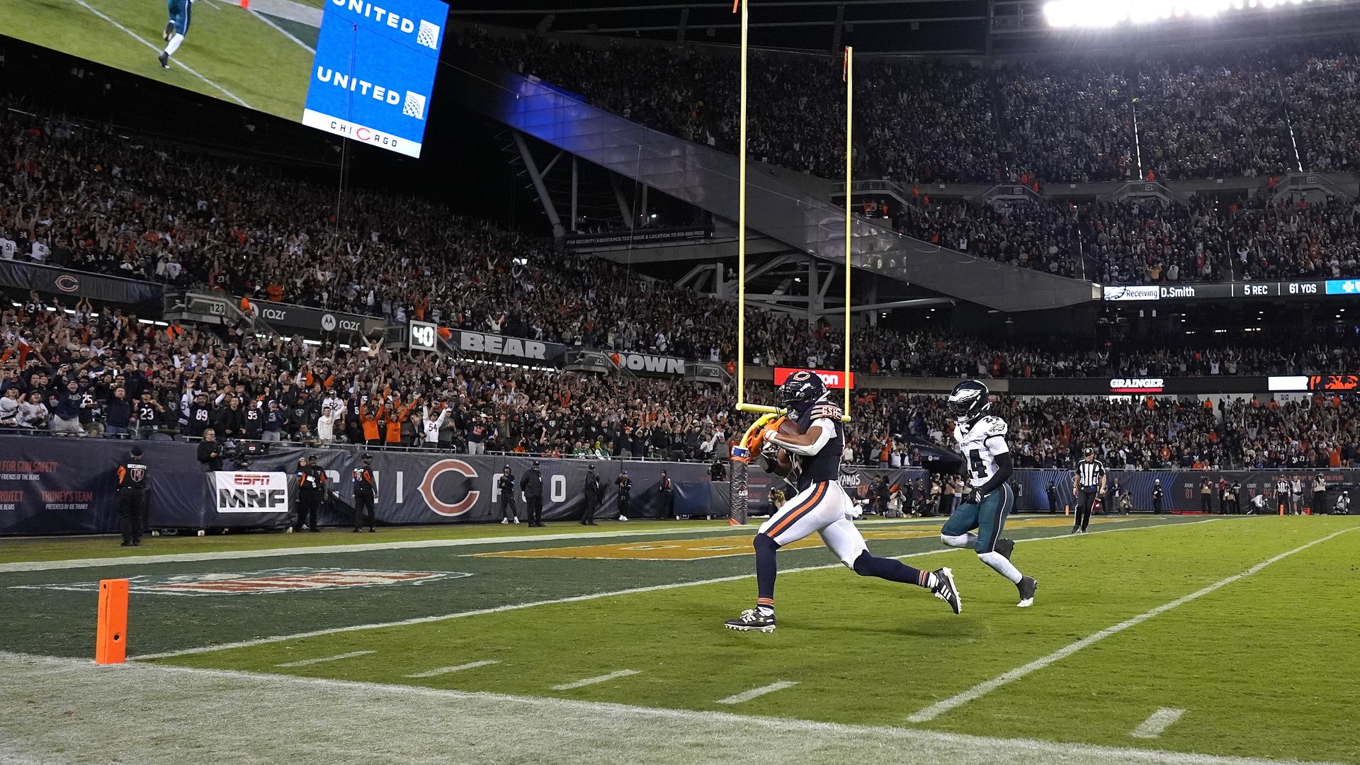 Night football game in a full stadium. Bears in navy and orange near the end zone chase a white/teal opponent; a bright blue "UNITED" scoreboard and goalposts loom as the crowd cheers.