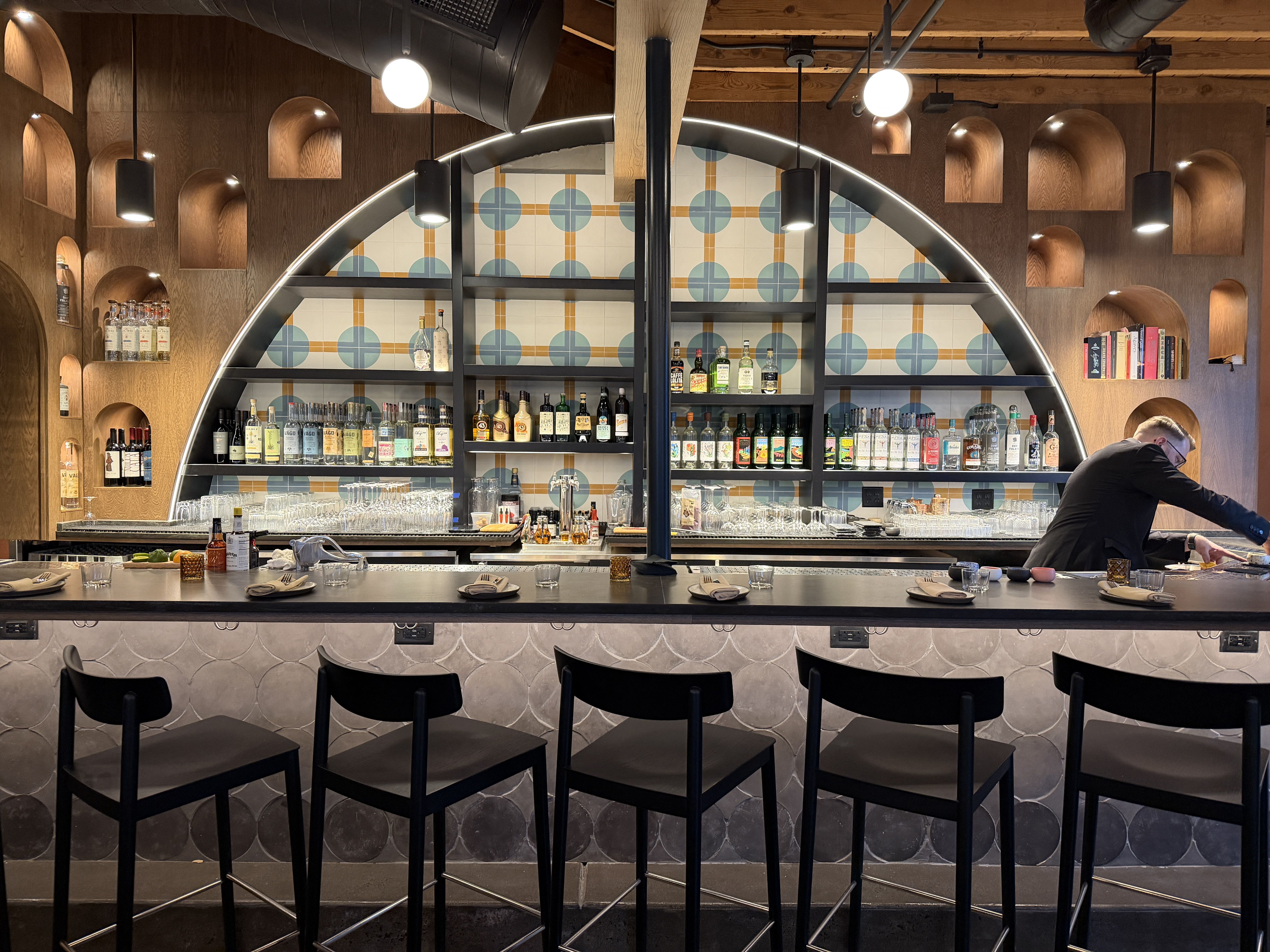 Modern bar with wooden wall featuring curved shelves and blue-orange tiled backdrop, black bar stools, dim lighting, and a person arranging items on the right side behind the counter.