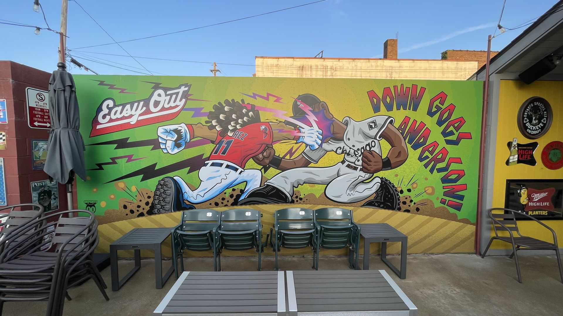Colorful mural of two cartoon baseball players fighting on a bright green wall: a red-jersey hitter labeled \"Rez 11\" and a white Chicago pitcher, with the text \"Easy Out\" and \"Down Goes\" on the wall; outdoor seating in front.