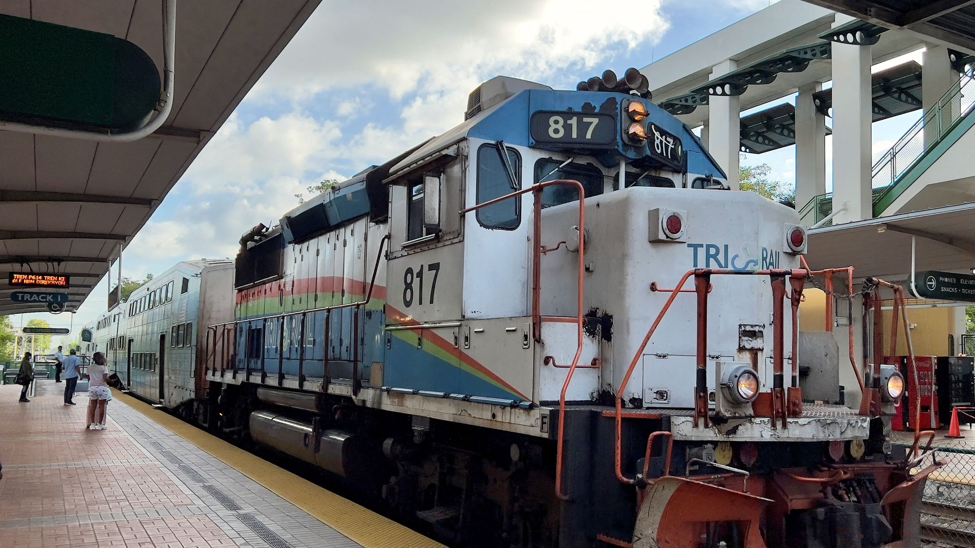 Blue and white TRI-RAIL train number 817 stopped at a sunny outdoor station platform with passengers waiting nearby under a partly cloudy sky.
