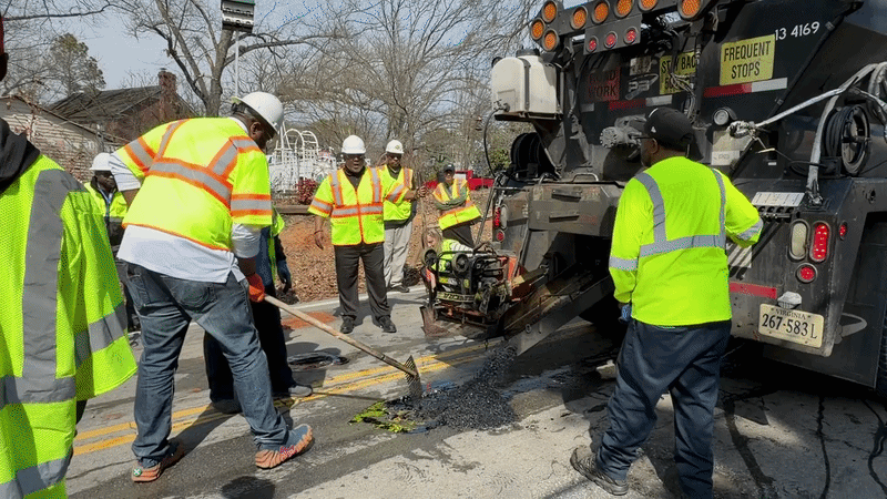 A gif showing a man in a bright construction vest filling a pothole with asphalt by raking it over the pothole