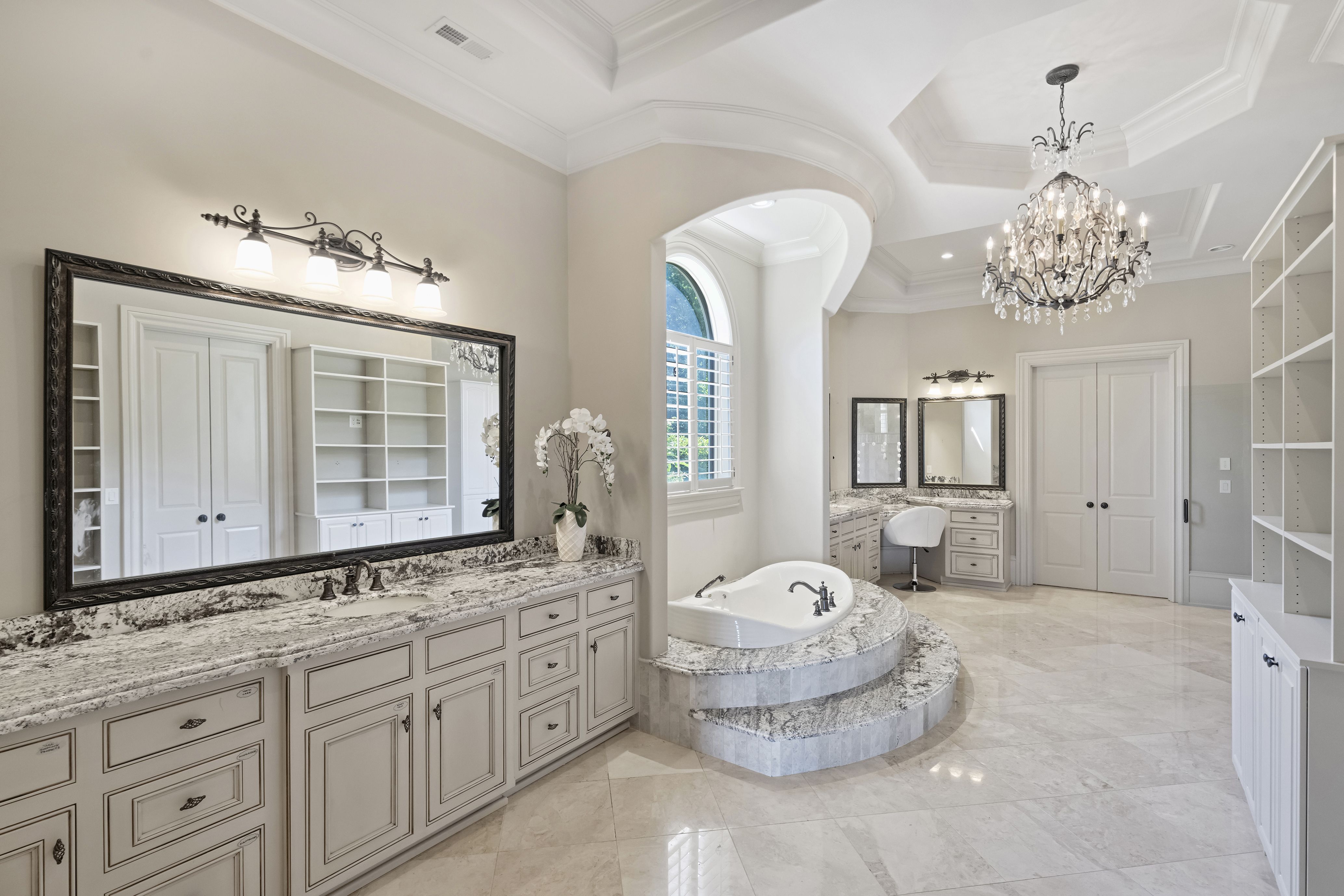 Luxurious bathroom with marble countertops and cabinetry, a large ornate framed mirror, four vanity lights, a crystal chandelier, arched window, and white built-in shelves.