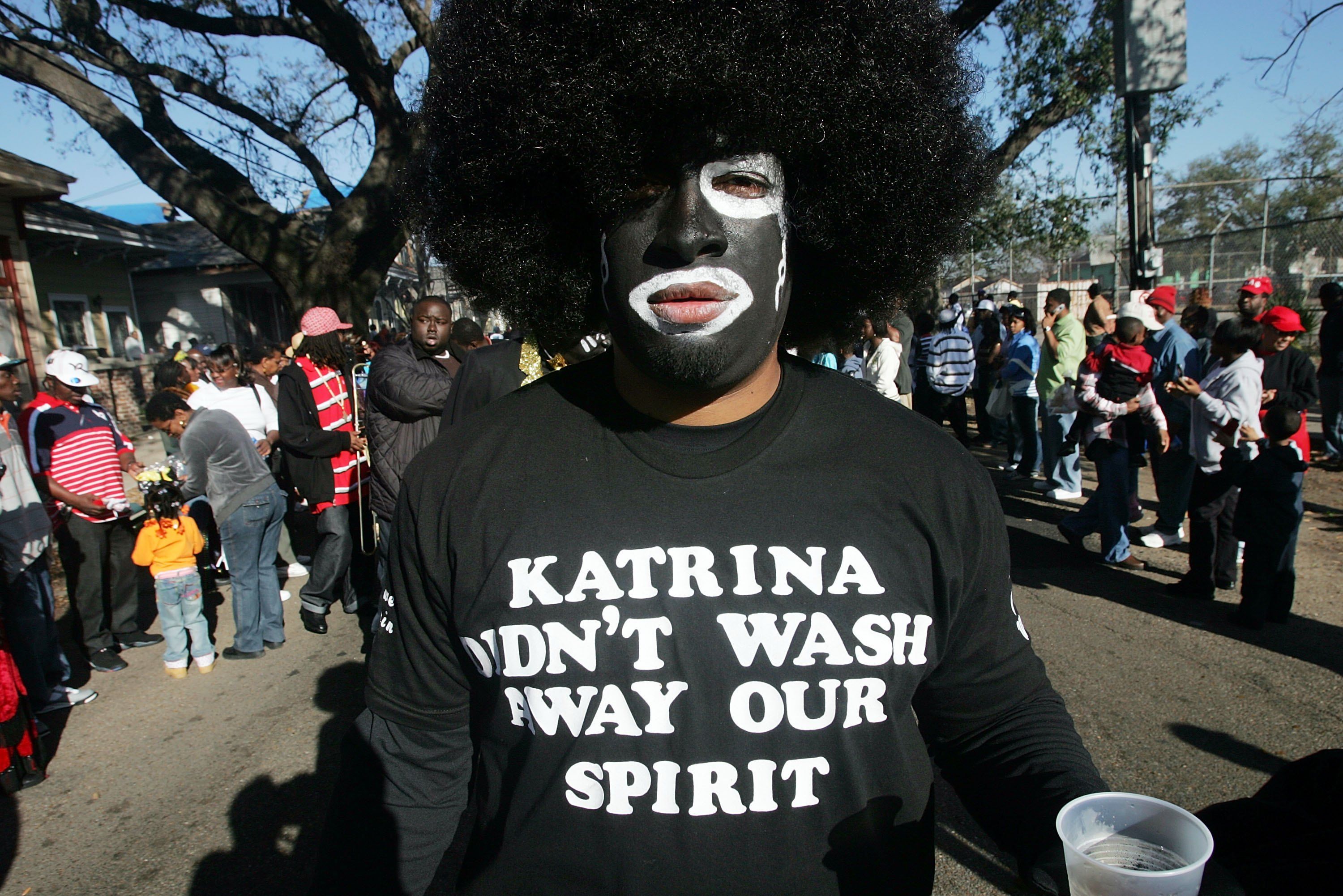 Person with an afro wig and face painted black and white wears a black shirt reading "Katrina didn't wash away our spirit" among a crowd on a sunny day outdoors.