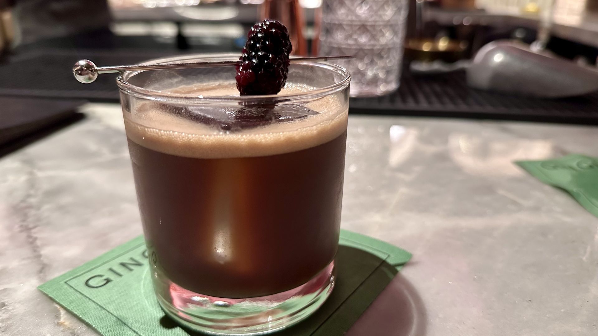 Dark brown cocktail in a clear glass with foam on top, garnished with a blackberry on a metal pick, sitting on a green coaster on a marble surface.