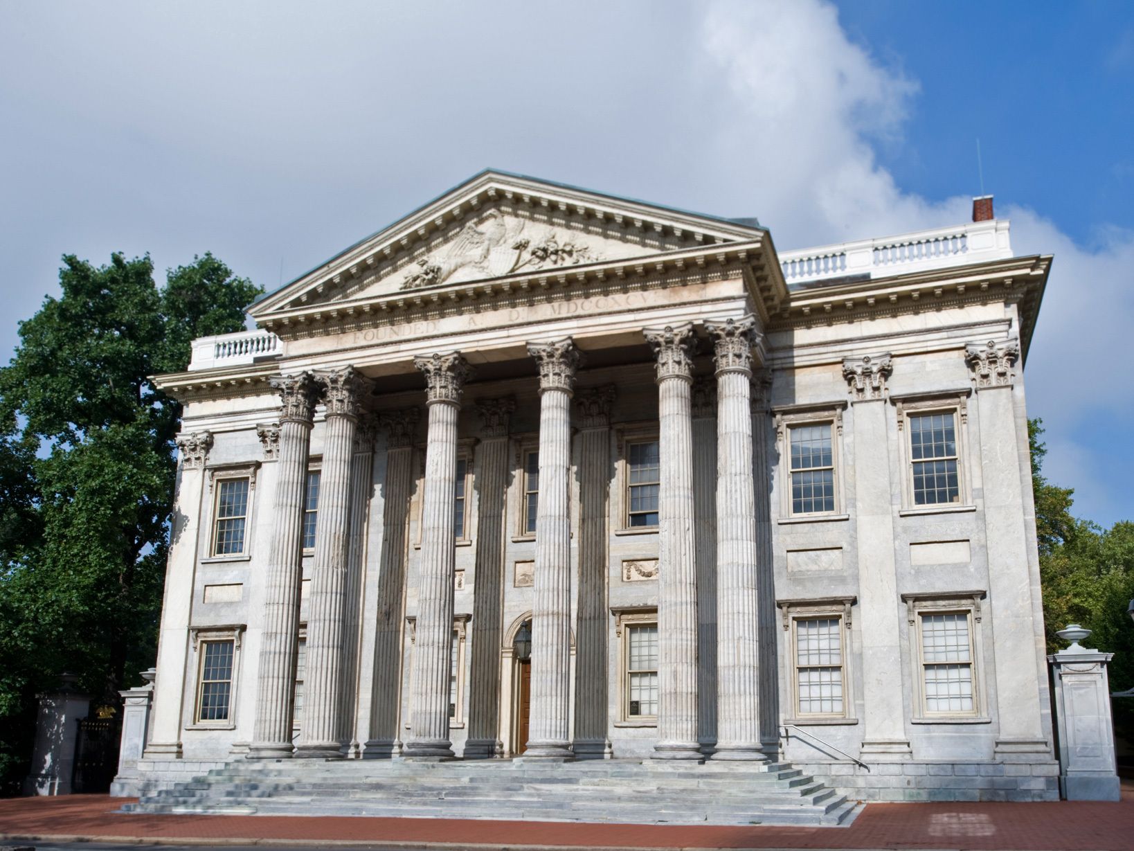 Neoclassical stone building with tall Corinthian columns, a triangular pediment, and wide steps. Surrounded by trees under a blue sky with clouds.