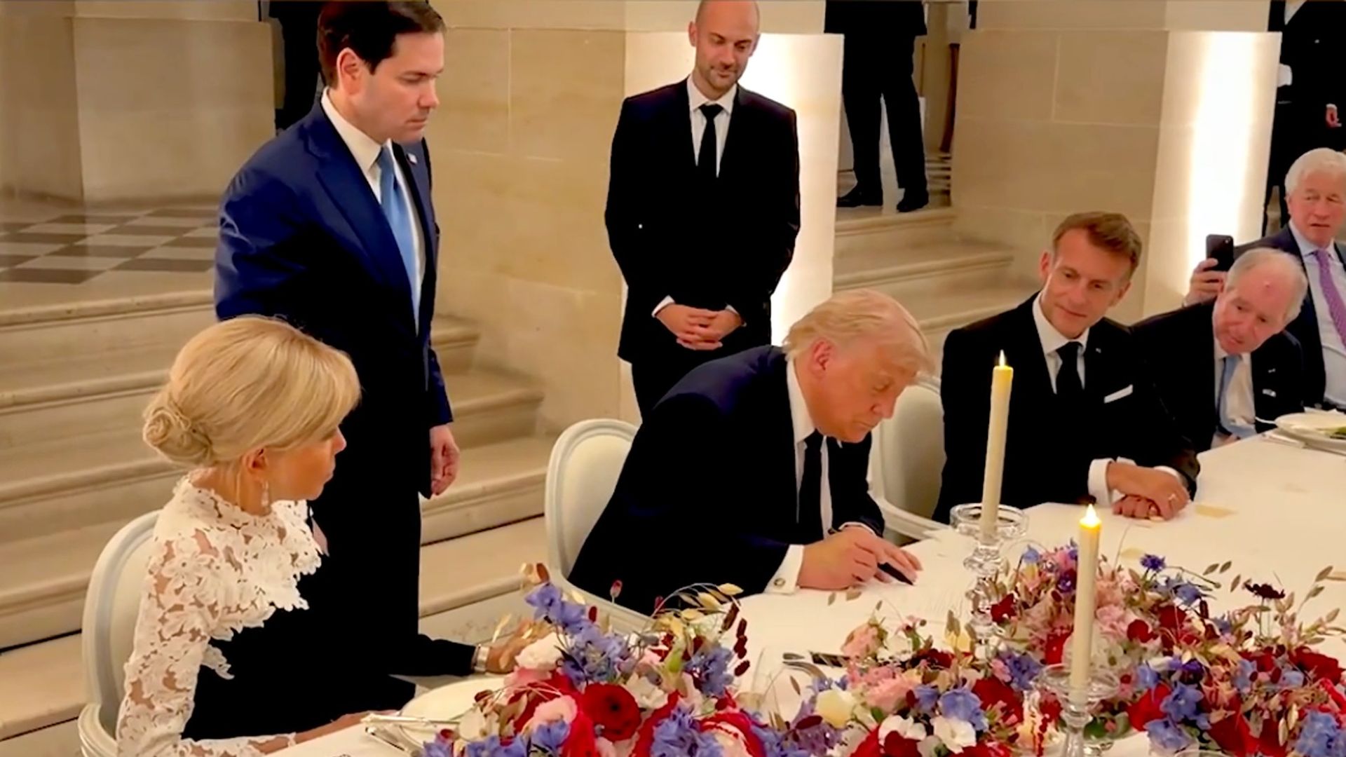 Formal dinner: a man in a dark suit signs a document at a long table with candles and a floral centerpiece; a blonde woman in white lace sits left; others in suits stand nearby in a hall.