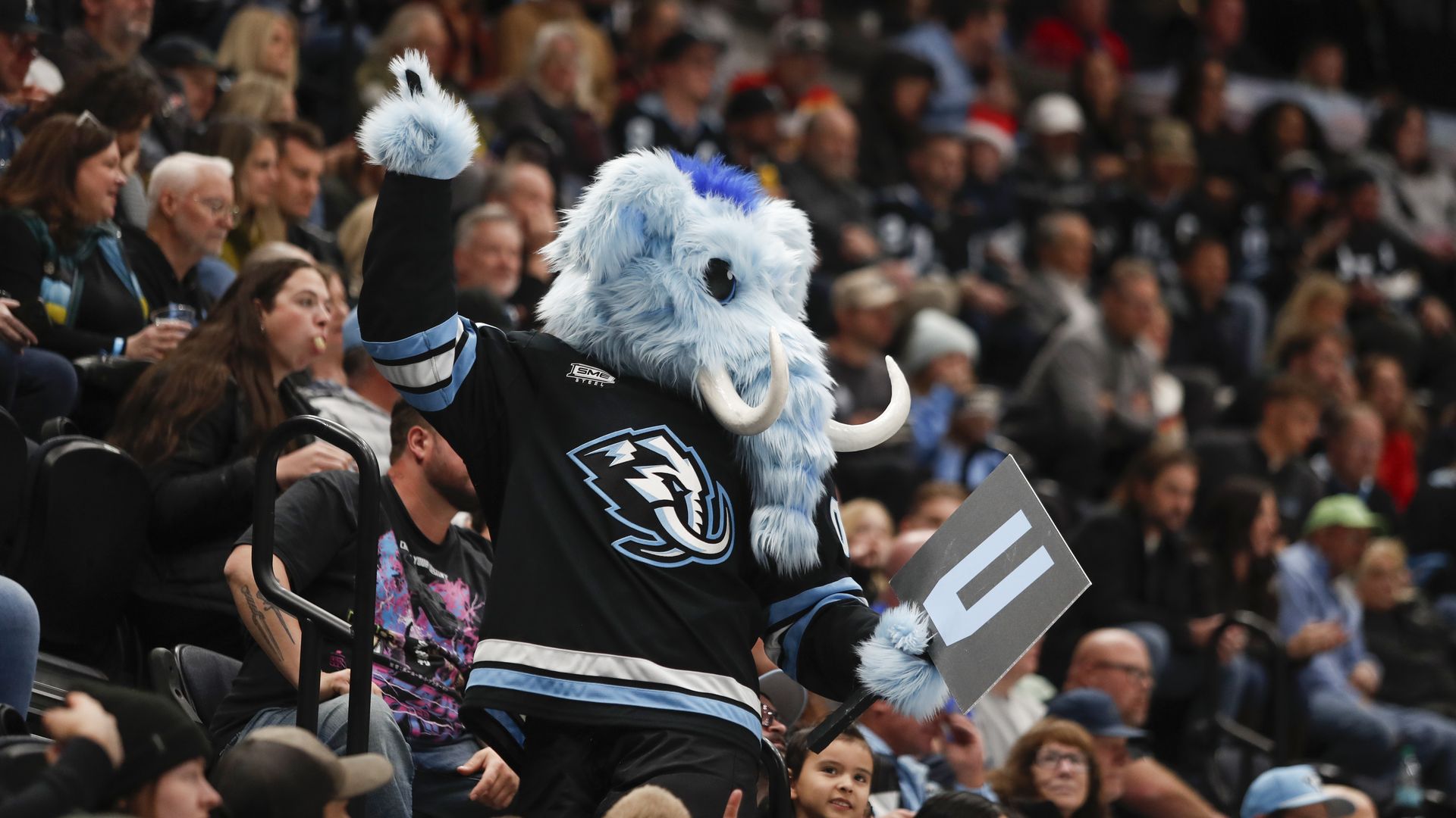 Blue fluffy mammoth mascot in a black and blue hockey jersey raises its right arm, waving to a crowded arena while holding a gray sign with blue/white markings; fans fill the stands behind.