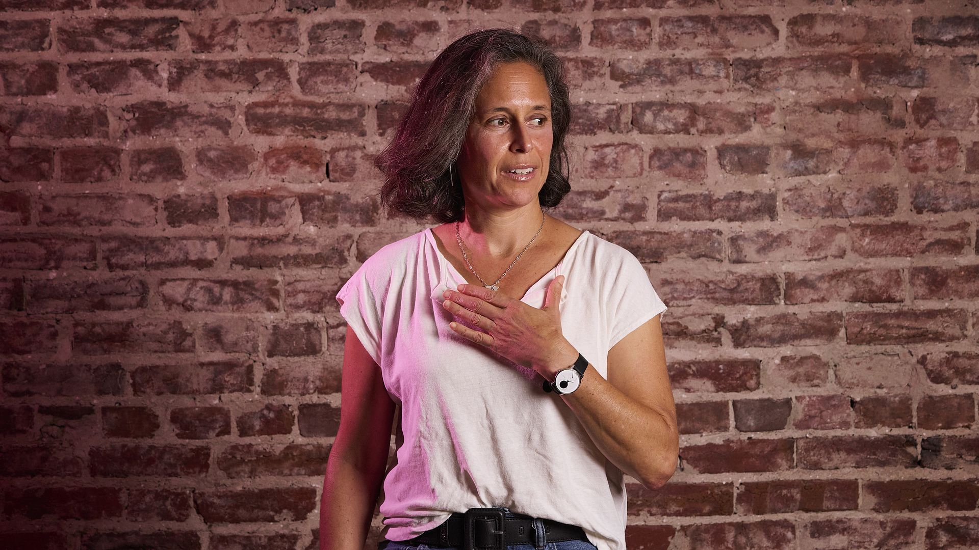 Woman with shoulder-length gray hair stands against a red brick wall, wearing a white T-shirt and jeans, with a necklace and watch. Her right hand rests on her chest as she looks to the right.