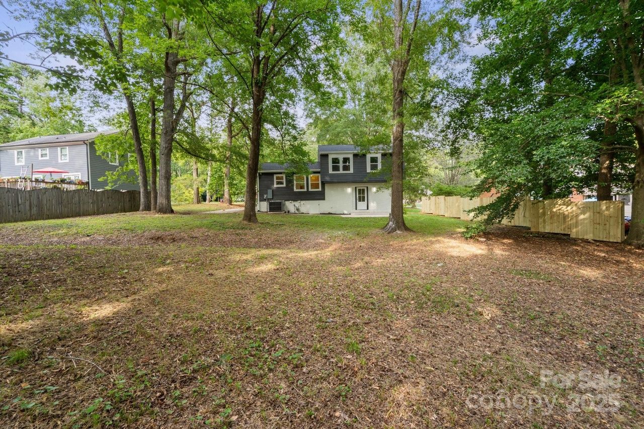 Backyard with sparse grass, scattered leaves, and several tall trees surrounding a two-story house with dark upper siding and white lower walls, under a clear sky.