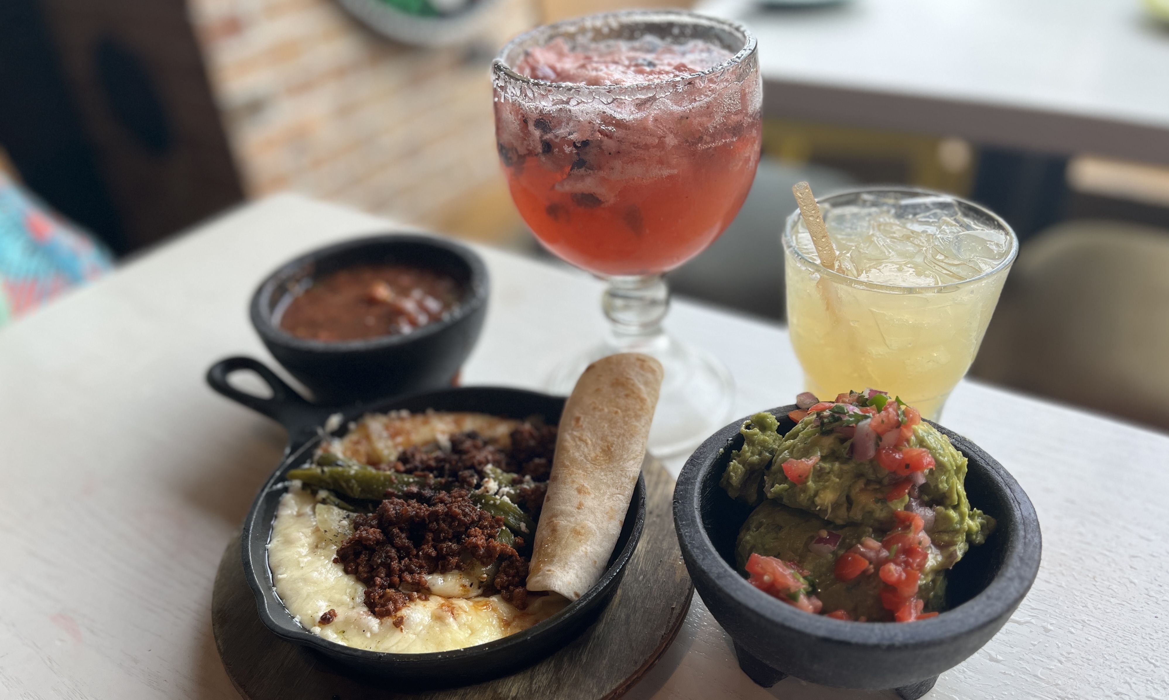 A table with Mexican dishes including queso fundido topped with chorizo and a flour tortilla, a bowl of guacamole with diced tomatoes, a bowl of salsa, a red drink with ice in a goblet glass, and a yellow drink with ice in a plastic cup.