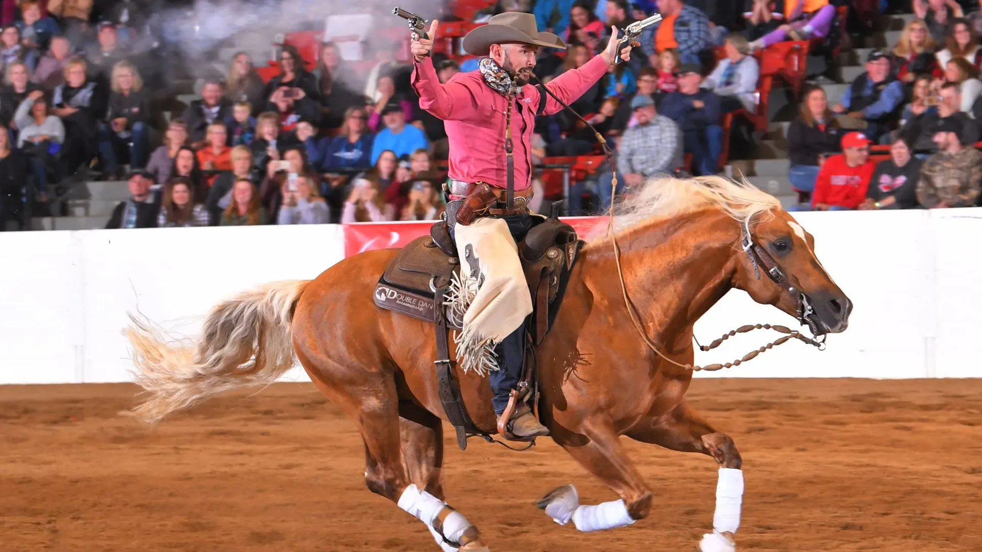 Cowboy in a pink shirt and tan hat rides a bucking brown horse in a dusty rodeo arena, arms raised with two pistols, fringe chaps, crowd cheering behind a white barrier.