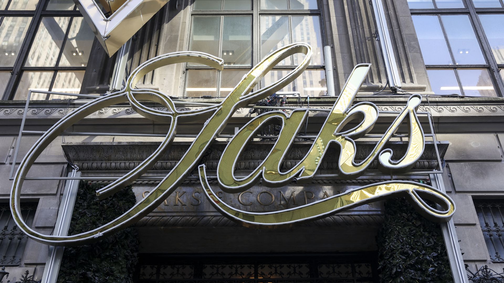 Large cursive golden sign reading "Saks" mounted on an ornate building facade with large windows and decorative stonework.