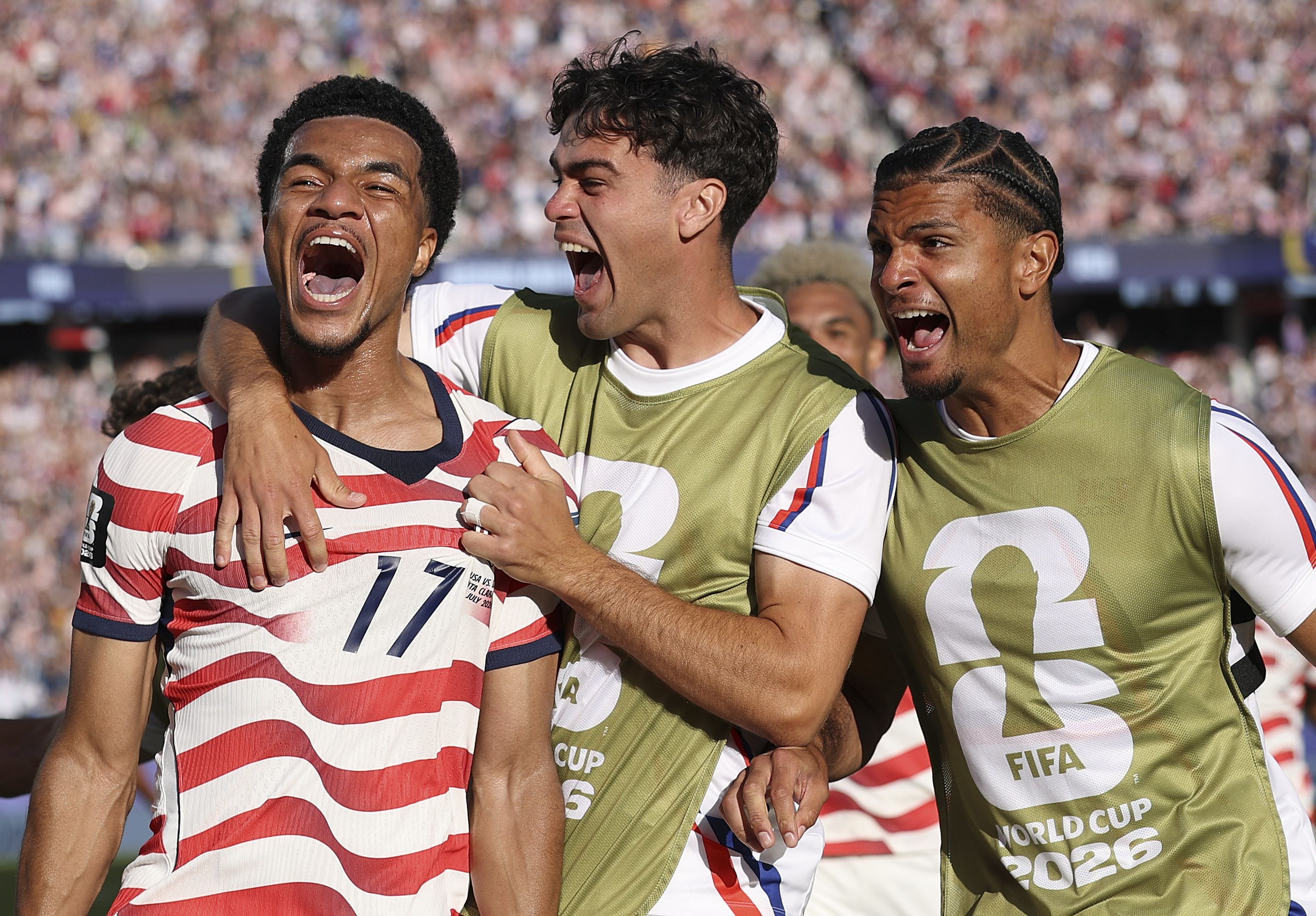 United States midfielder Malik Tillman shouts in celebration as teammates surround him after his goal during the FIFA World Cup Round of 32 match against Bosnia and Herzegovina in Santa Clara, California, with a packed stadium in the background.