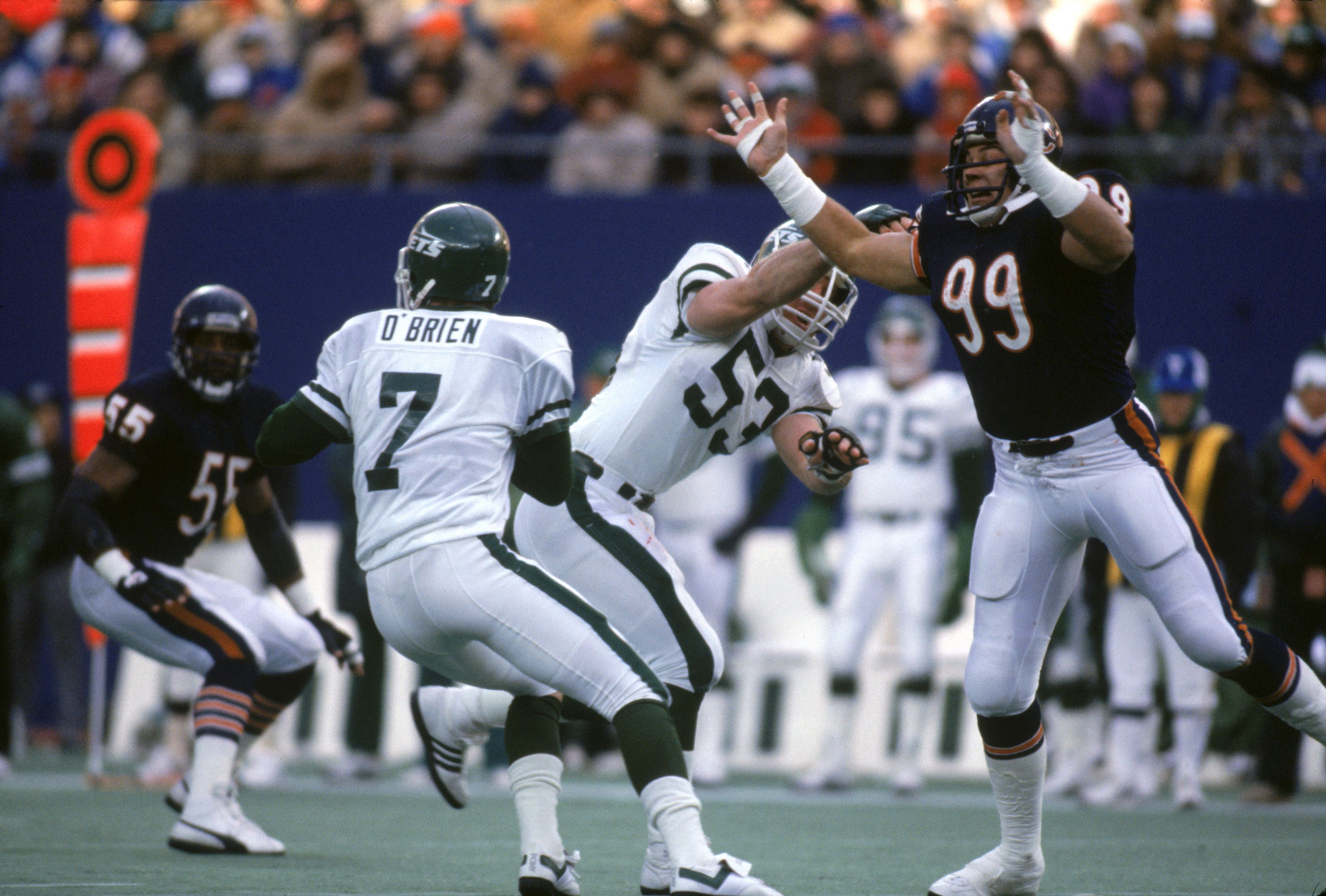 Football game action with a Jets player wearing number 7 named O'Brien preparing to pass as a defender in navy blue number 99 raises arms to block a white-uniformed player running. Crowd in background.