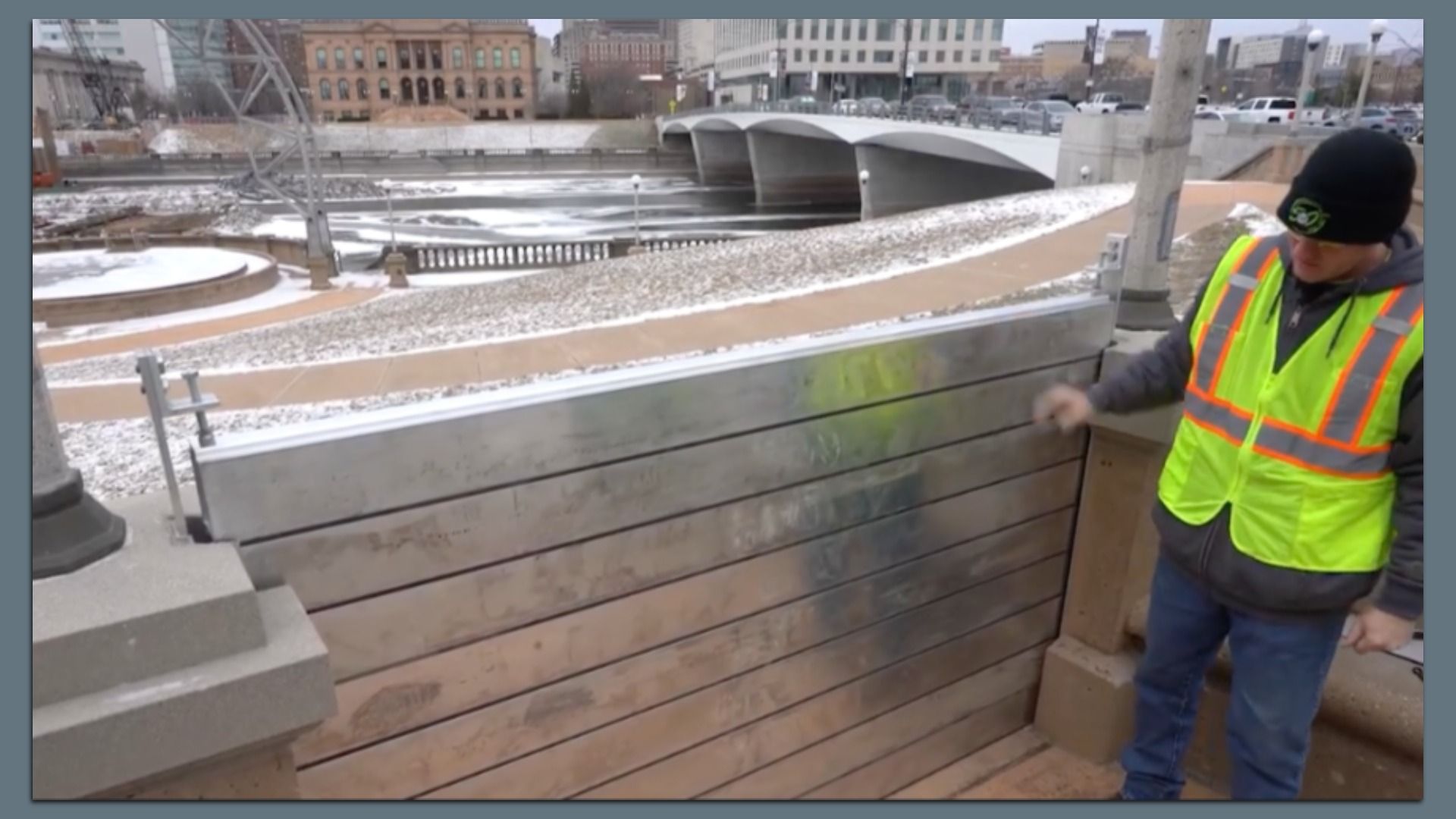 Man wearing a neon yellow safety vest and black beanie stands by a floodgate barrier in an urban area with snow dust on the ground and a concrete bridge over a partially frozen river in the background.