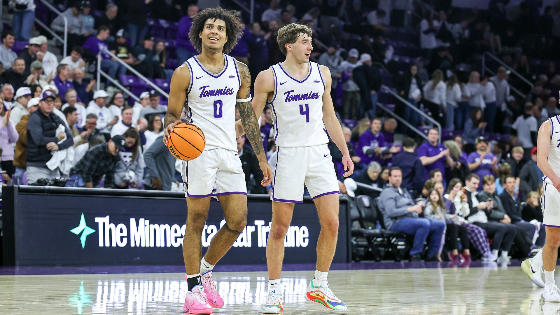 Two basketball players in white Tommies uniforms stand on a polished court; player 0 holds an orange ball, player 4 wears colorful shoes; crowd watches in background.