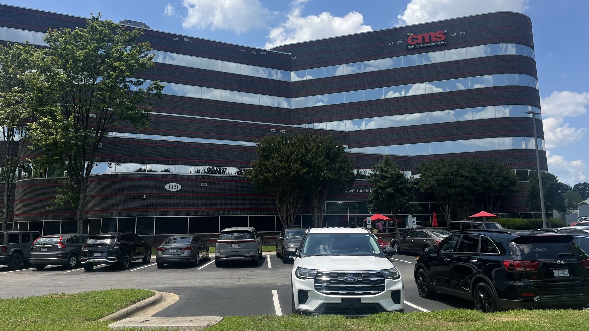 Black and red-brick multi-story office building with reflective windows under blue sky, parked cars in front, CMS logo for Charlotte-Mecklenburg Schools on upper right corner.