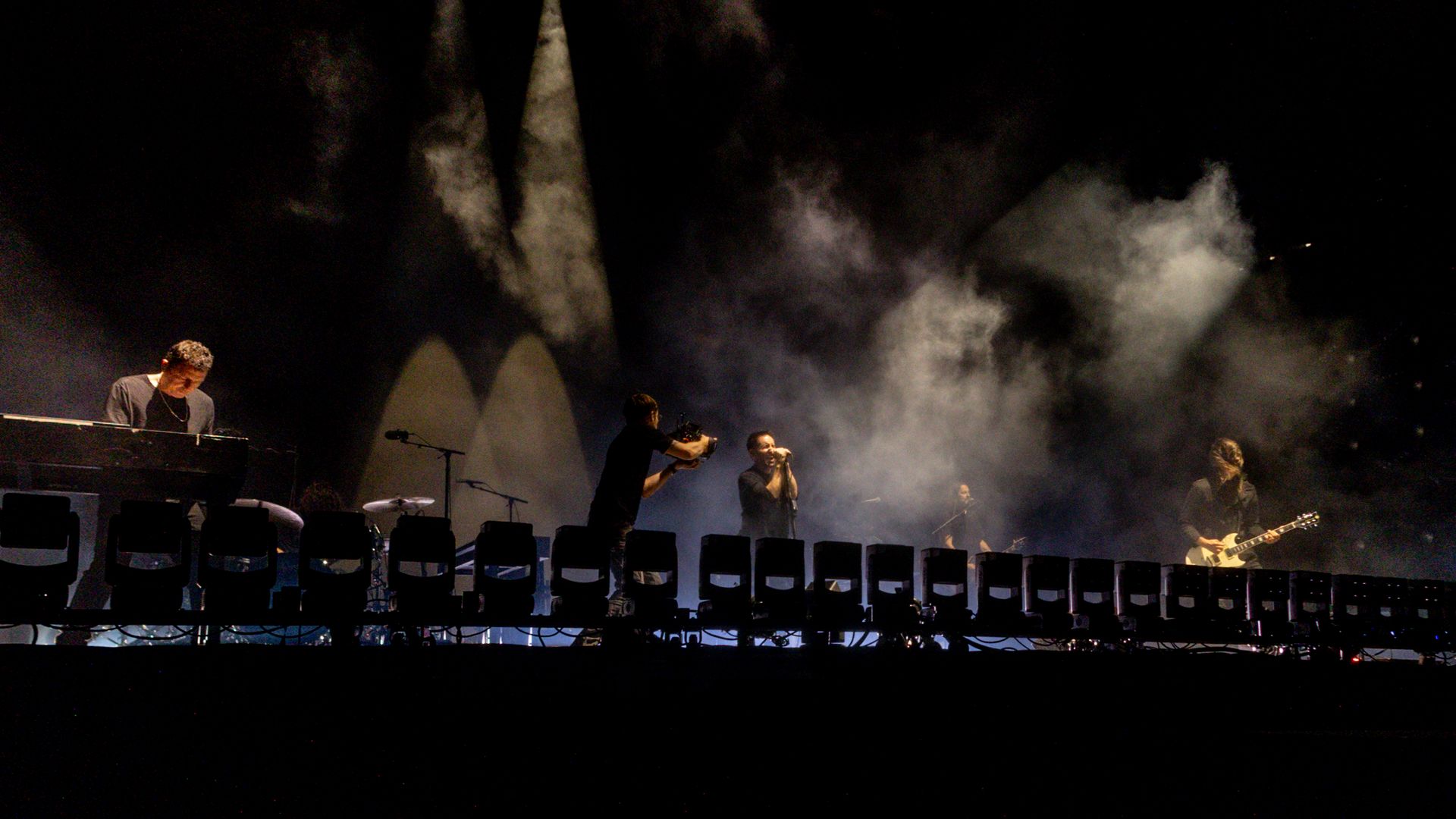A dark concert stage with musicians playing keyboard, guitar, and drums, a vocalist singing under spotlights with smoke effects, and a cameraman filming the performance.