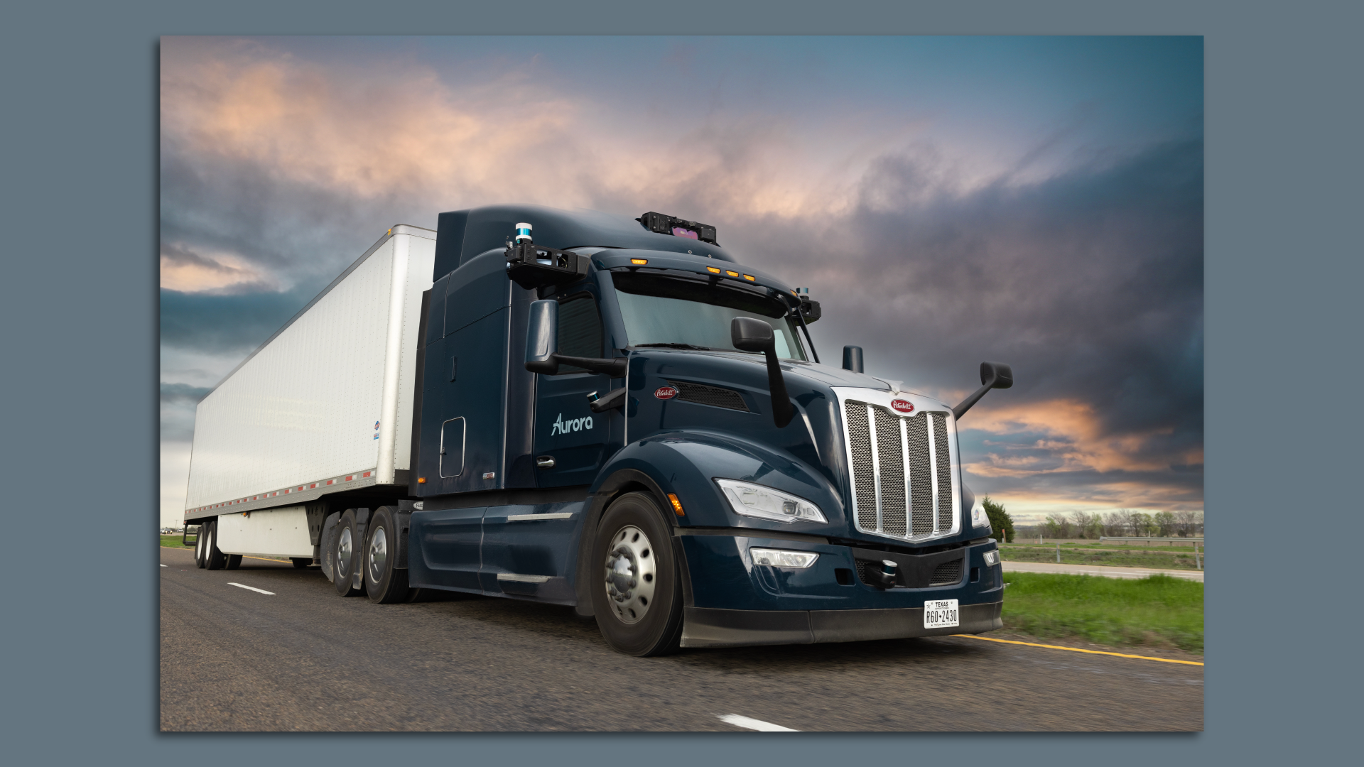Dark blue autonomous semi-truck with "Aurora" logo on the door, hauling a white trailer on a highway under a dramatic cloudy sky at sunset.