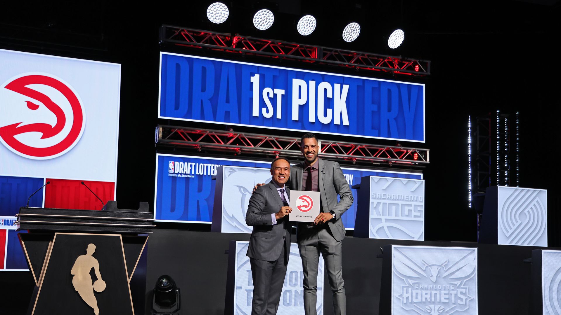Two men stand in front of a sign that says 1st pick holding the Atlanta Hawks logo