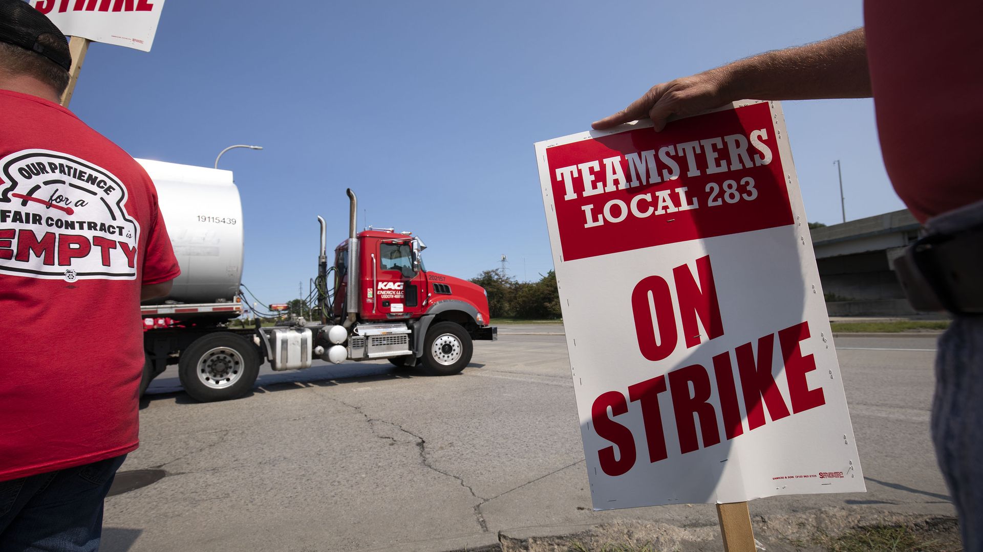 Two people are visible holding strike signs and wearing red shirts with a truck in the background 