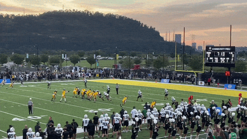 High school football game showing running back runing 20 yards into the end zone with green hills and orange sky in background.