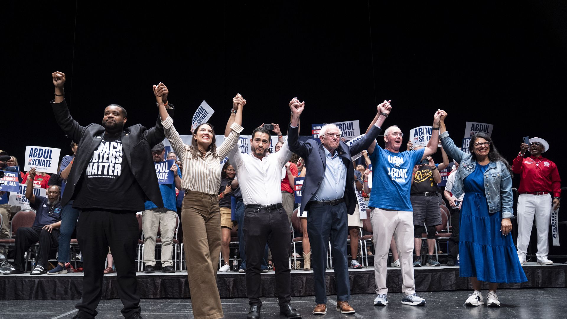Abdul El-Sayed stands onstage with Alexandria Ocasio-Cortez, Bernie Sanders, Shawn Fain, Rashida Tlaib and Donavan McKinney, raising joined hands before a crowd of supporters holding campaign signs in Detroit.