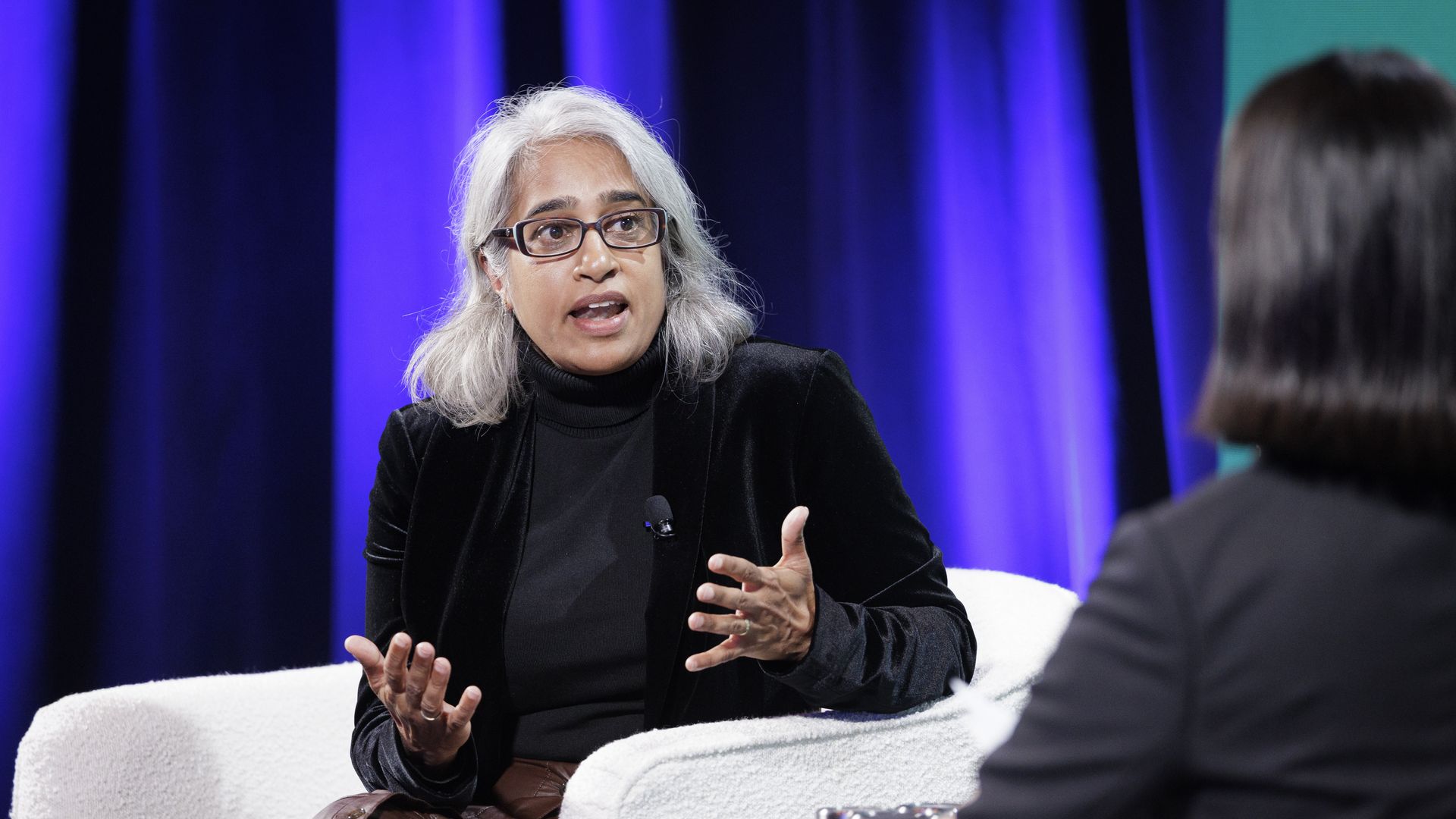 Two women in a discussion on stage with purple lighting, one with white hair in black jacket and brown skirt speaking expressively while seated in a white chair, the other in black suit seen from behind.