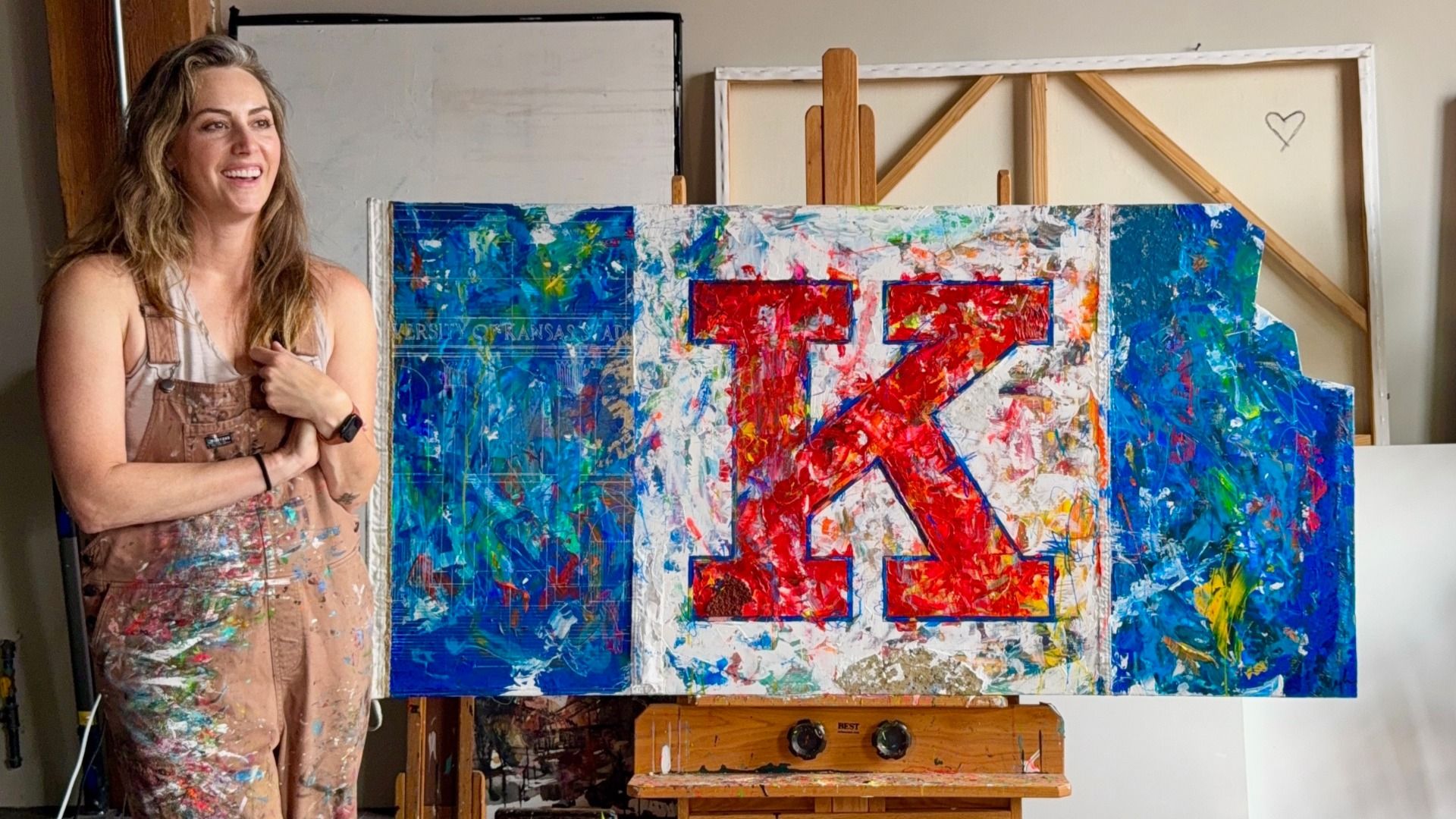 Smiling woman in paint-splattered brown overalls stands next to colorful abstract painting with large red letter K on white and blue background, in an art studio.