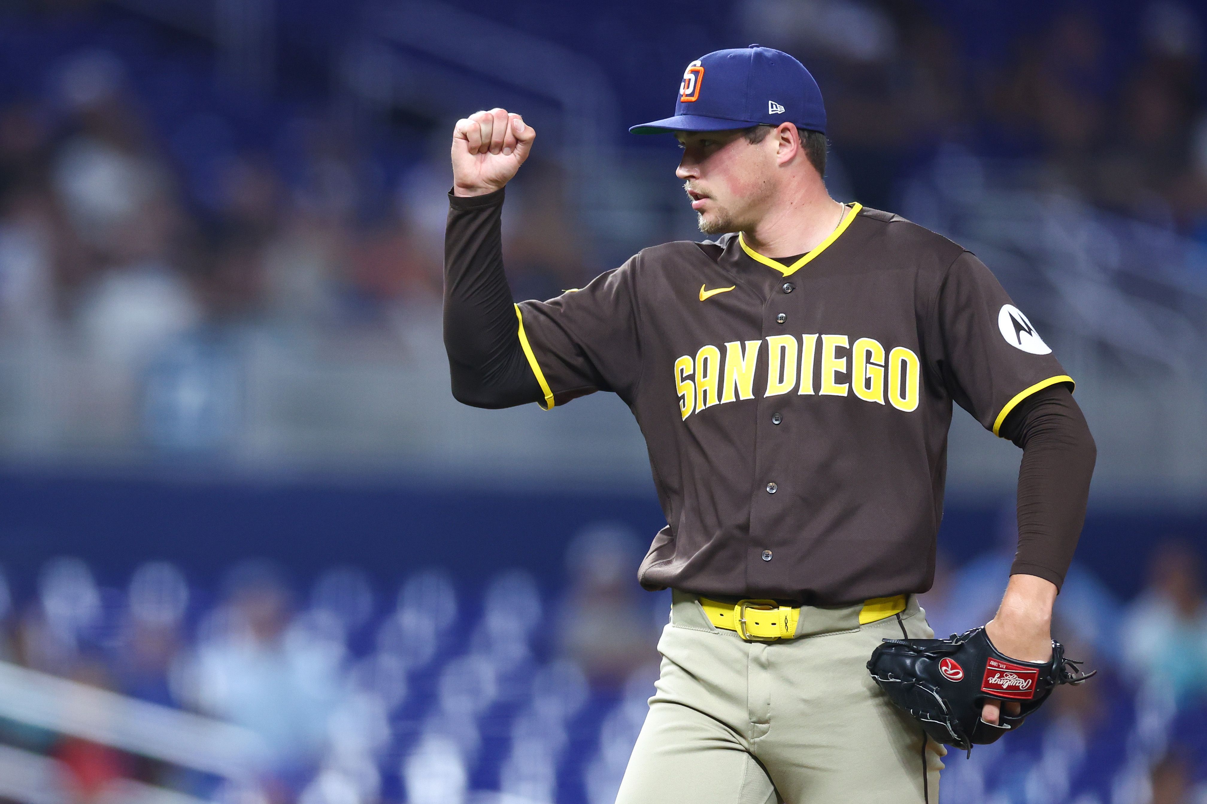 A San Diego Padres pitcher in brown jersey with yellow trim and blue cap raises a clenched fist on the mound, glove in hand, blurred stadium seats in the background.