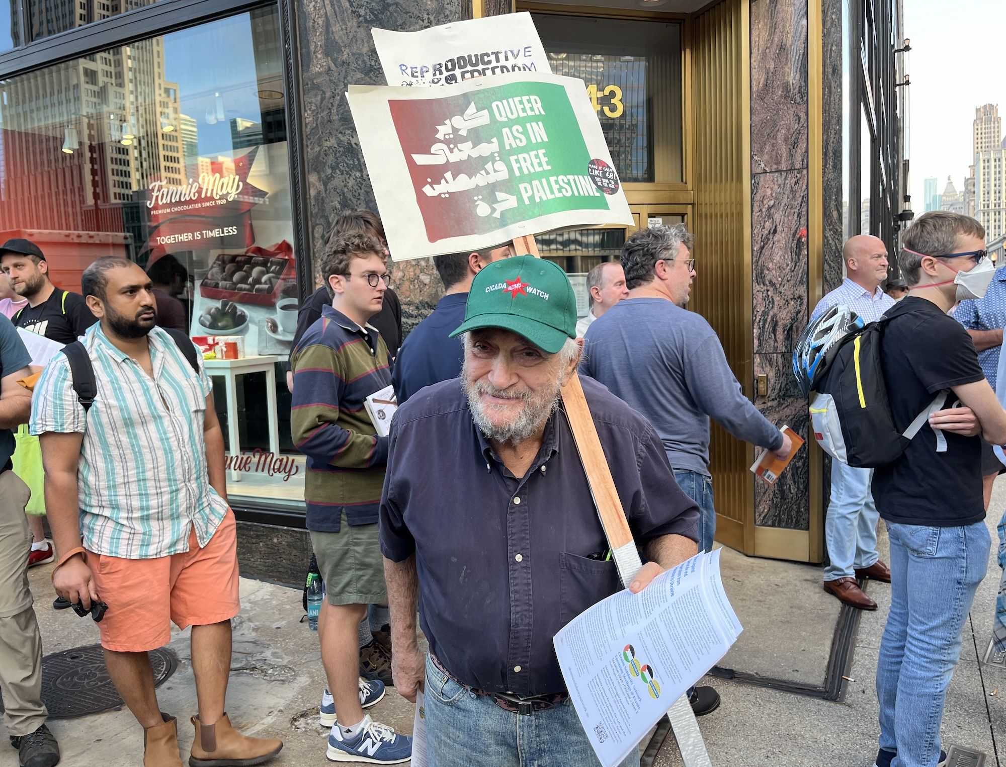 Man in green hate holding a sign that reads Queer as in Free Palestine.