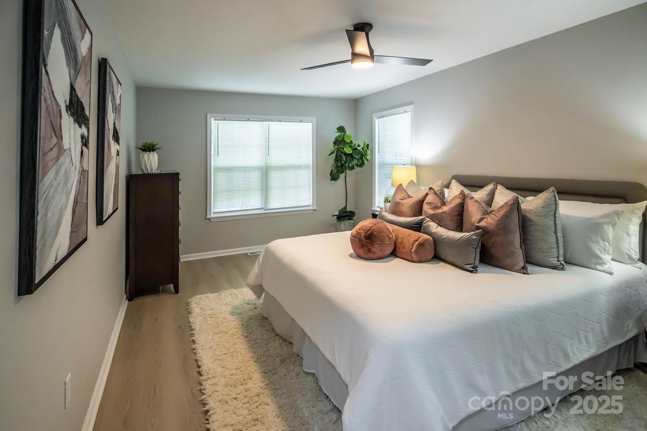 Modern bedroom with a large bed featuring white bedding and various brown and gray pillows, light wood floor, white shag rug, abstract artwork on gray walls, and a ceiling fan.