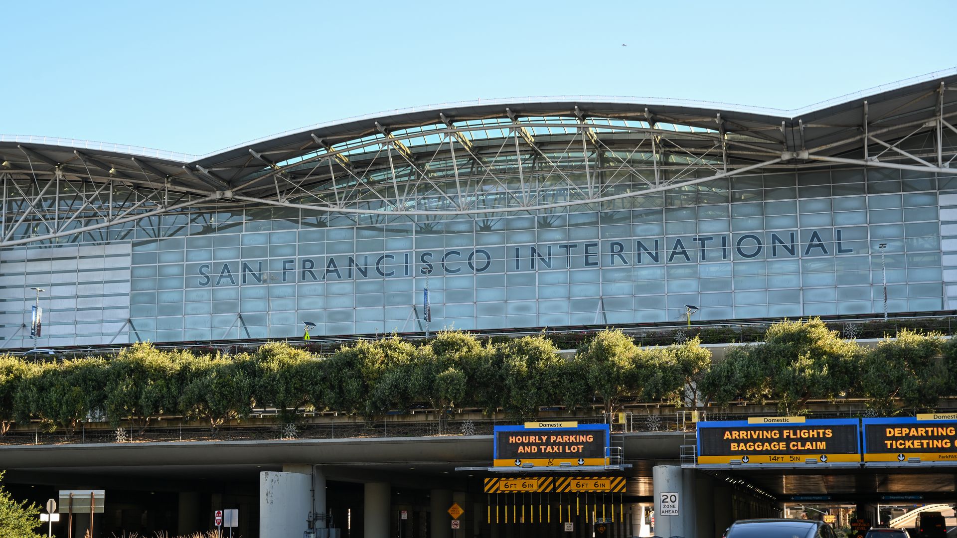 View of San Francisco International Airport terminal with glass façade and metal roof. A large SAN FRANCISCO INTERNATIONAL sign; cars drive past digital signs for parking, arrivals, and departures.