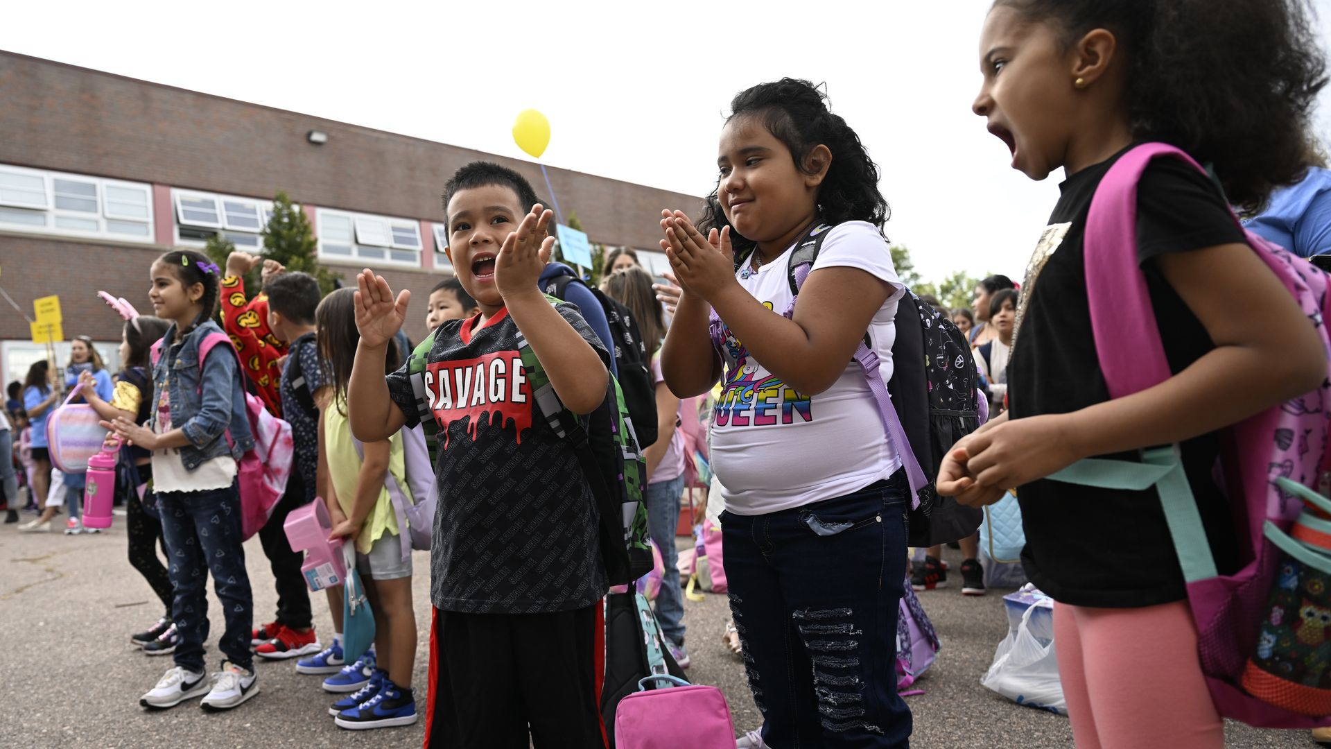 Three children in the foreground clap and sing while other young children stand behind them during a sunny day outside an elementary school in Denver.