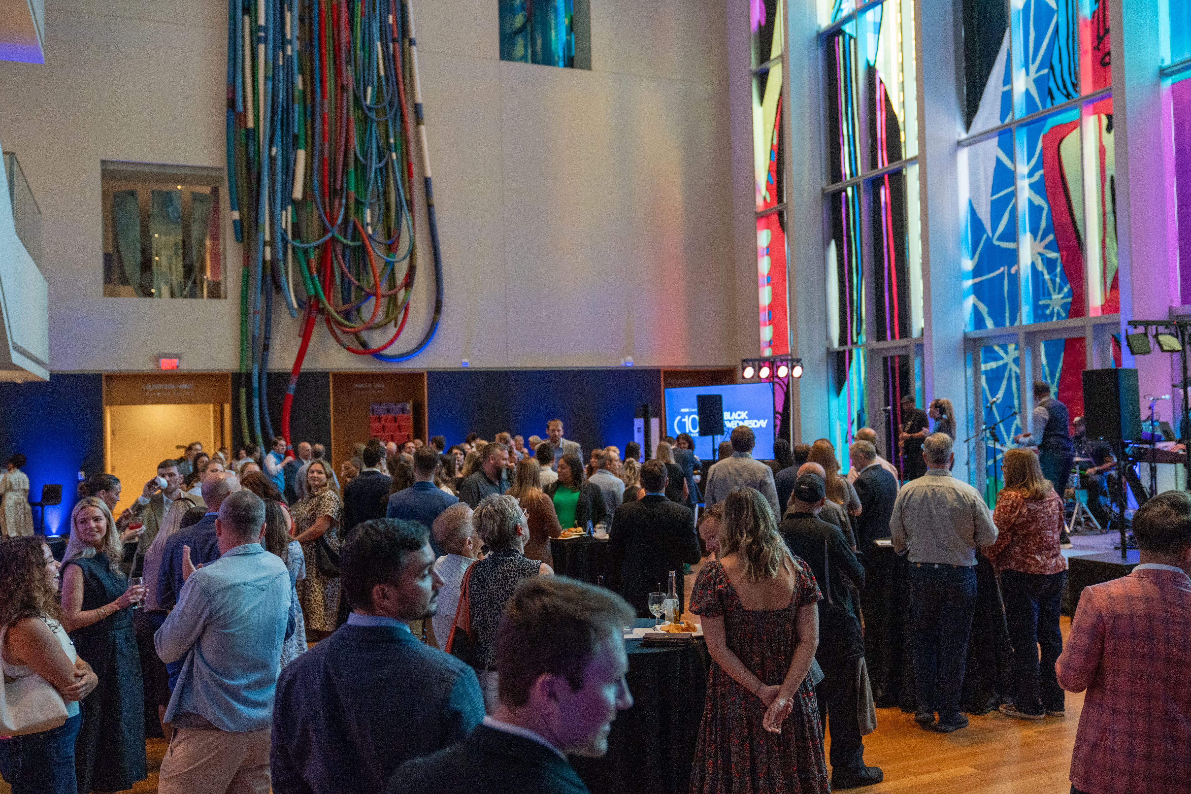 Crowded indoor event with people dressed in casual to semi-formal attire, colorful abstract wall art, and a screen displaying "Black Wednesday" in a brightly lit room.