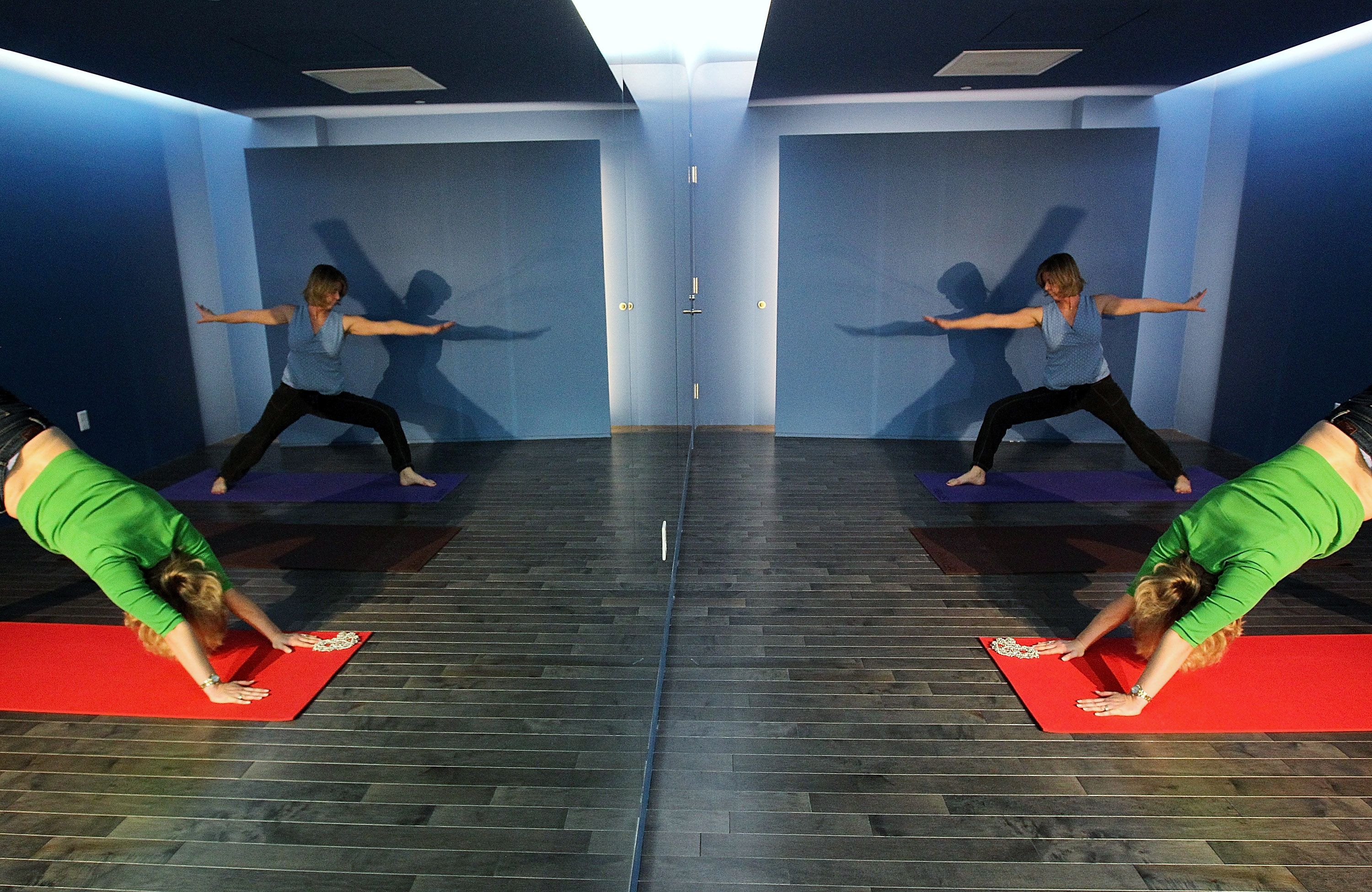 Two women in a blue room with dark wood floors doing yoga on mats; one in green on a red mat in downward dog pose, the other in blue on purple mat in a warrior pose, reflected in a mirror.