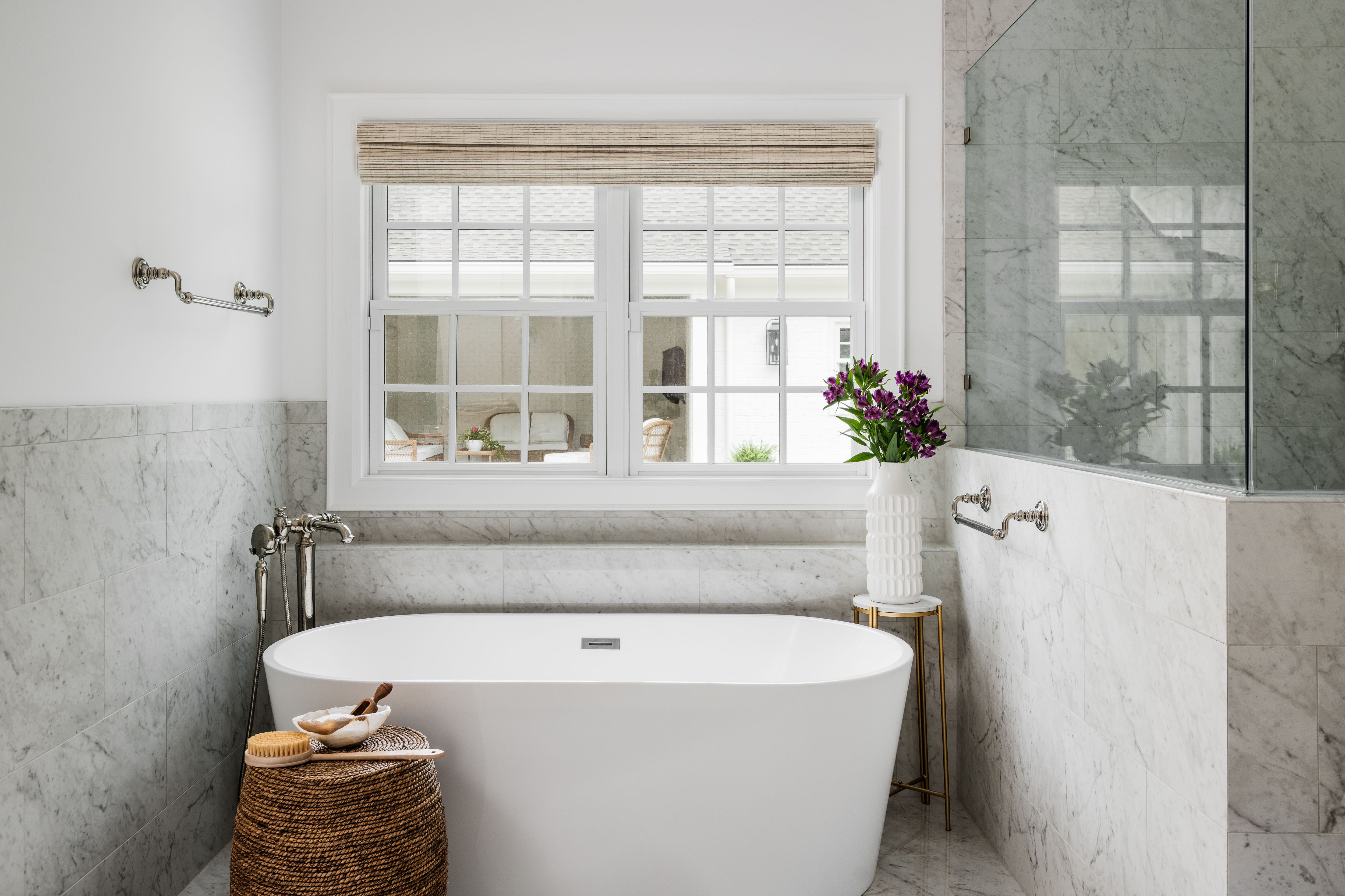 Bright bathroom with white freestanding bathtub, marble walls, wicker stool holding bath items, window with beige blind, and white vase with purple flowers on gold stand.