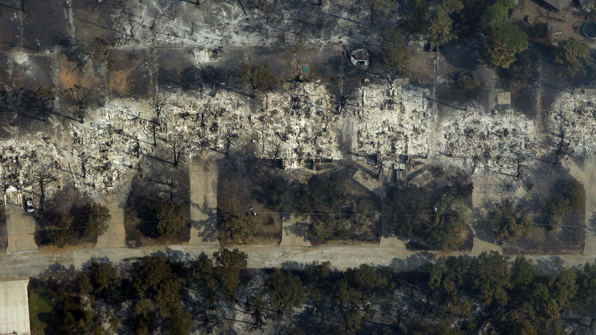 An aerial view of a neighborhood after a wildfire: a rubble-strewn strip with charred trees and scorched ground along a road, green lawns nearby, smoke fading in the distance.