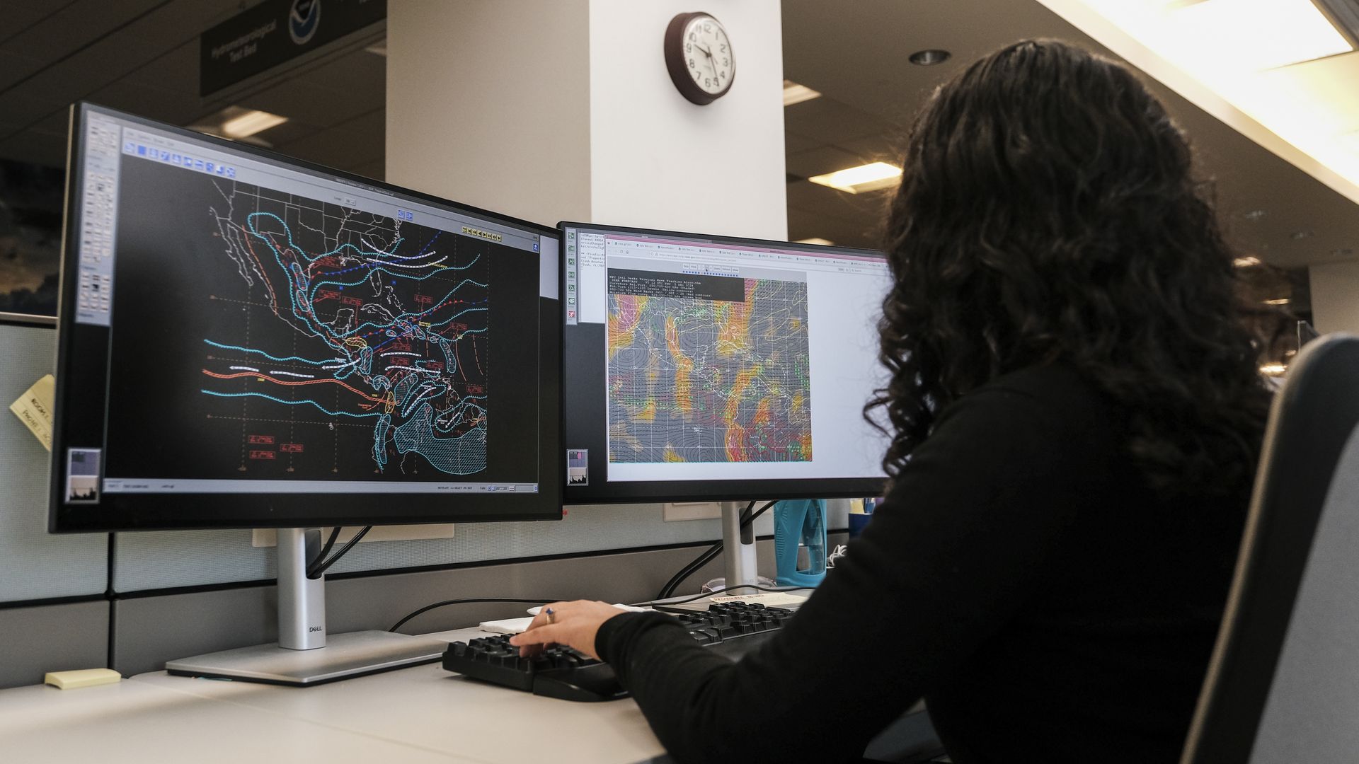 A woman sits at a desk looking at two computer monitors that are displaying weather information on them.