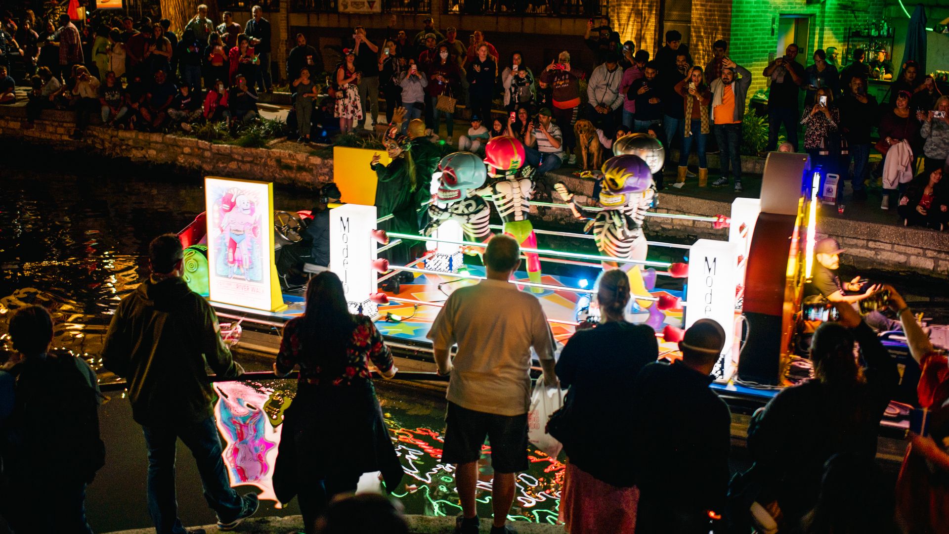 Nighttime riverside crowd watches a colorful skeleton boxing ring on a boat; spectators take photos as neon-lit brick buildings glow in green and warm lights.