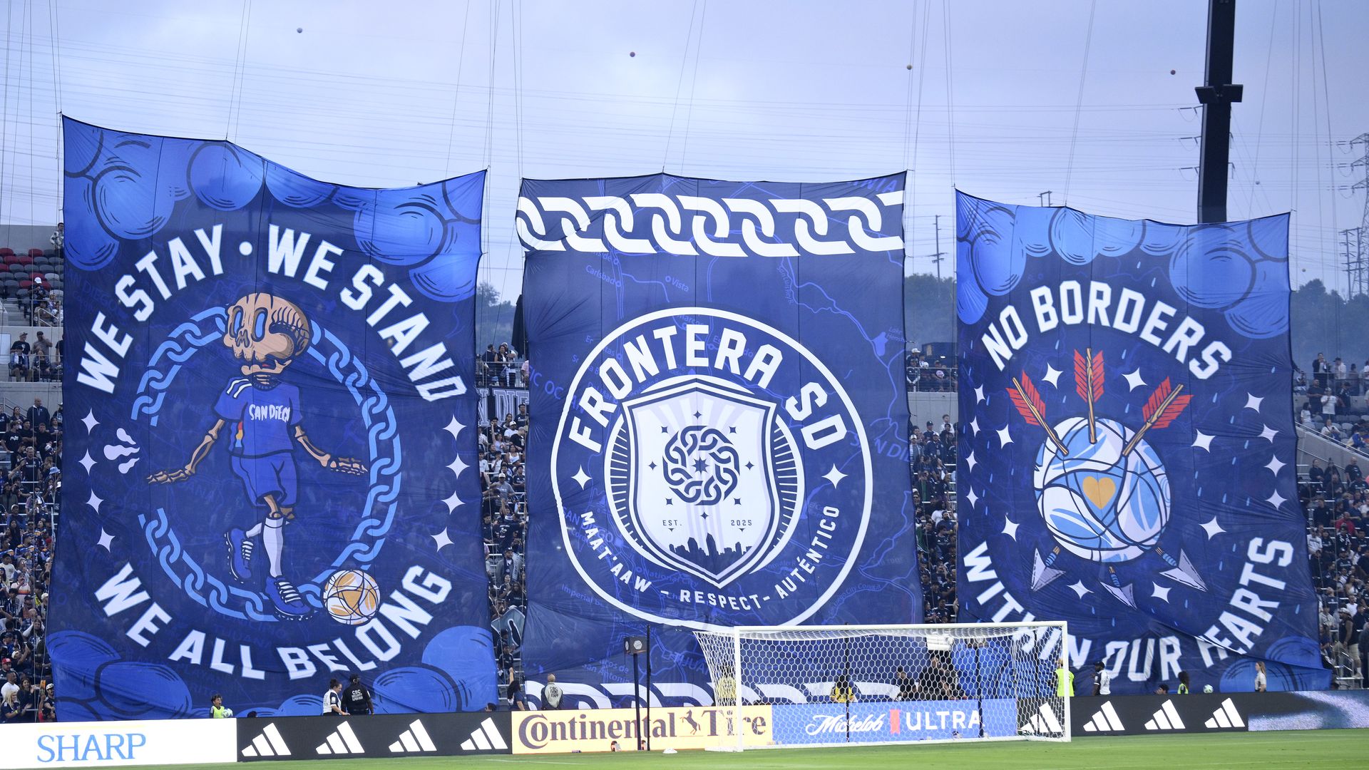 Three large blue banners at a soccer stadium with messages: "We stay, we stand, we all belong," "Frontera SD," and "No borders within our hearts," featuring soccer and symbolic images.