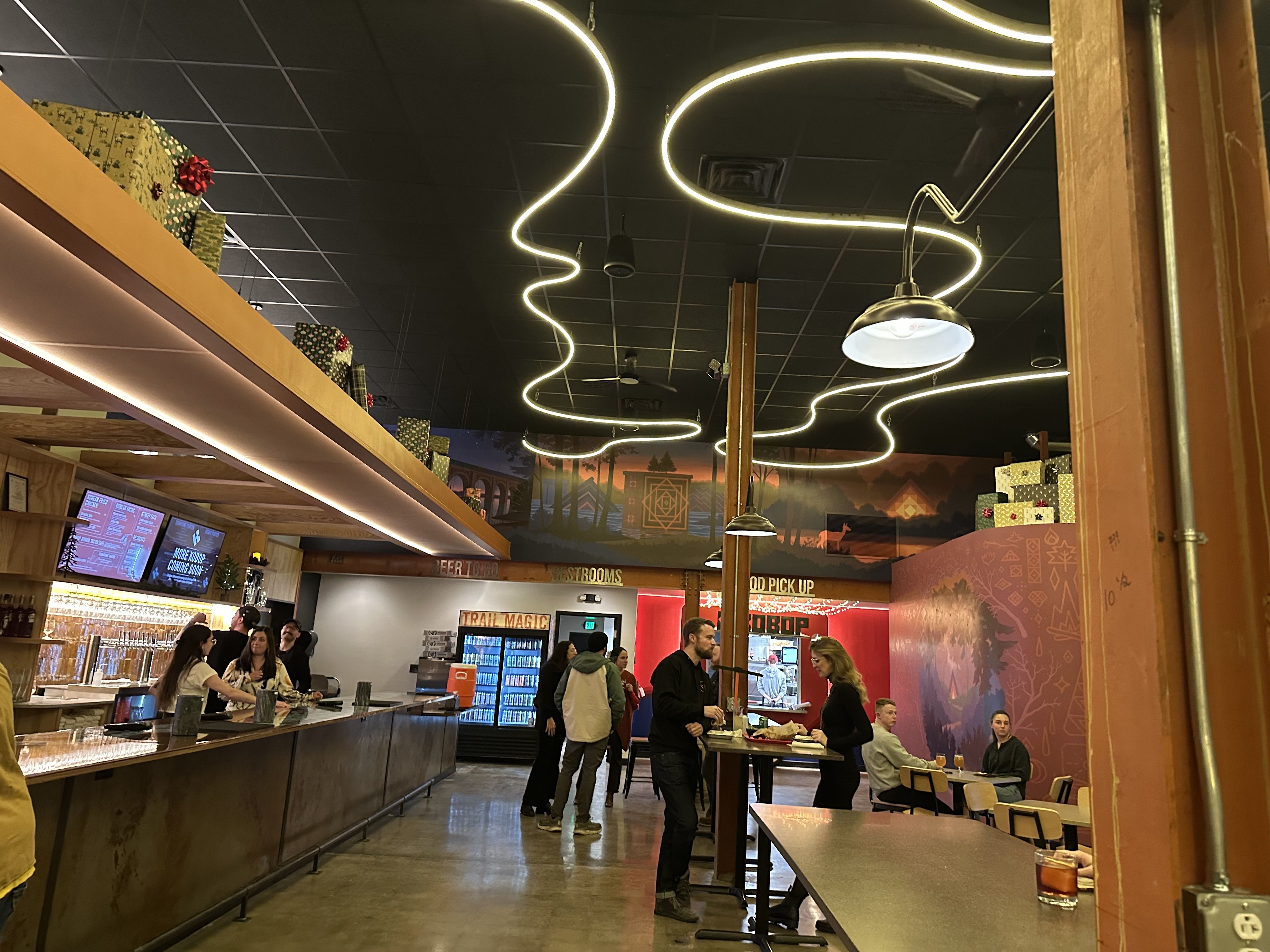 Inside a cafe with glowing neon squiggle lights on a dark ceiling, a long counter on the left, gift boxes on a shelf, wooden beams, and customers gathered by a warm mural.