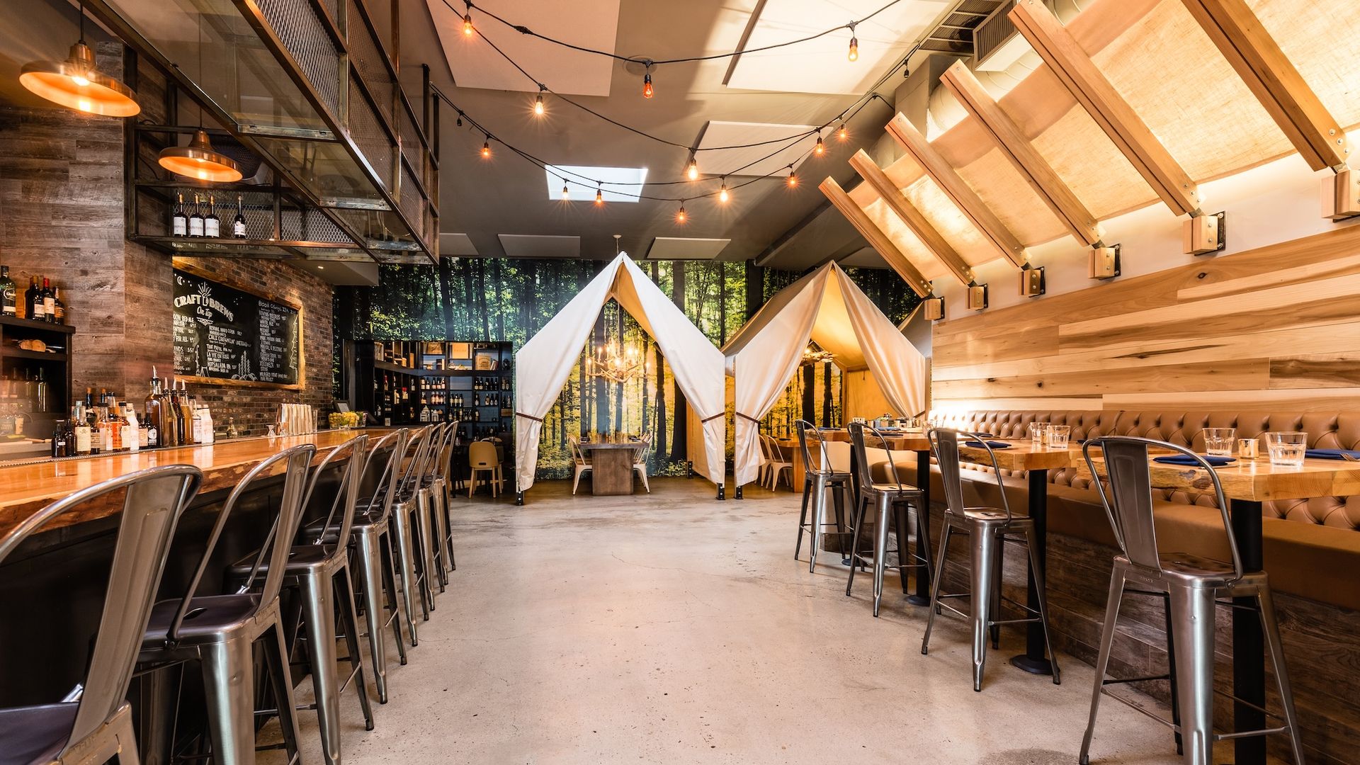 Modern, outdoorsy restaurant interior with metal bar stools, wooden tables, leather banquette seating, two white canvas tents, string lights overhead, and a forest mural on the back wall.